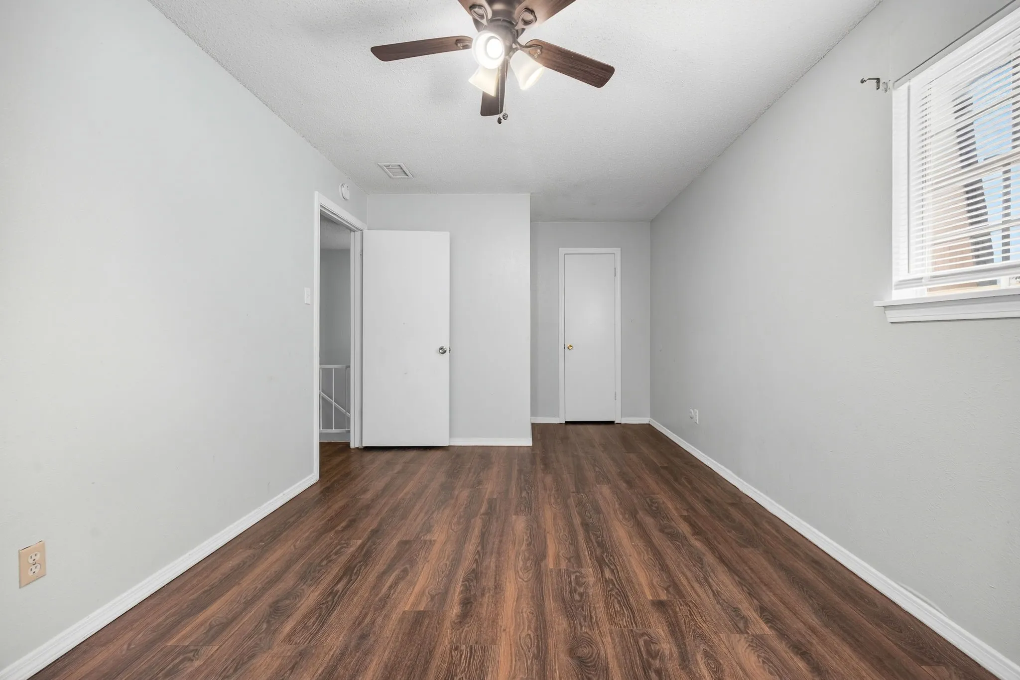 Spacious room featuring wood-finish flooring, light gray walls, a ceiling fan with integrated lighting, and a white-framed window with blinds