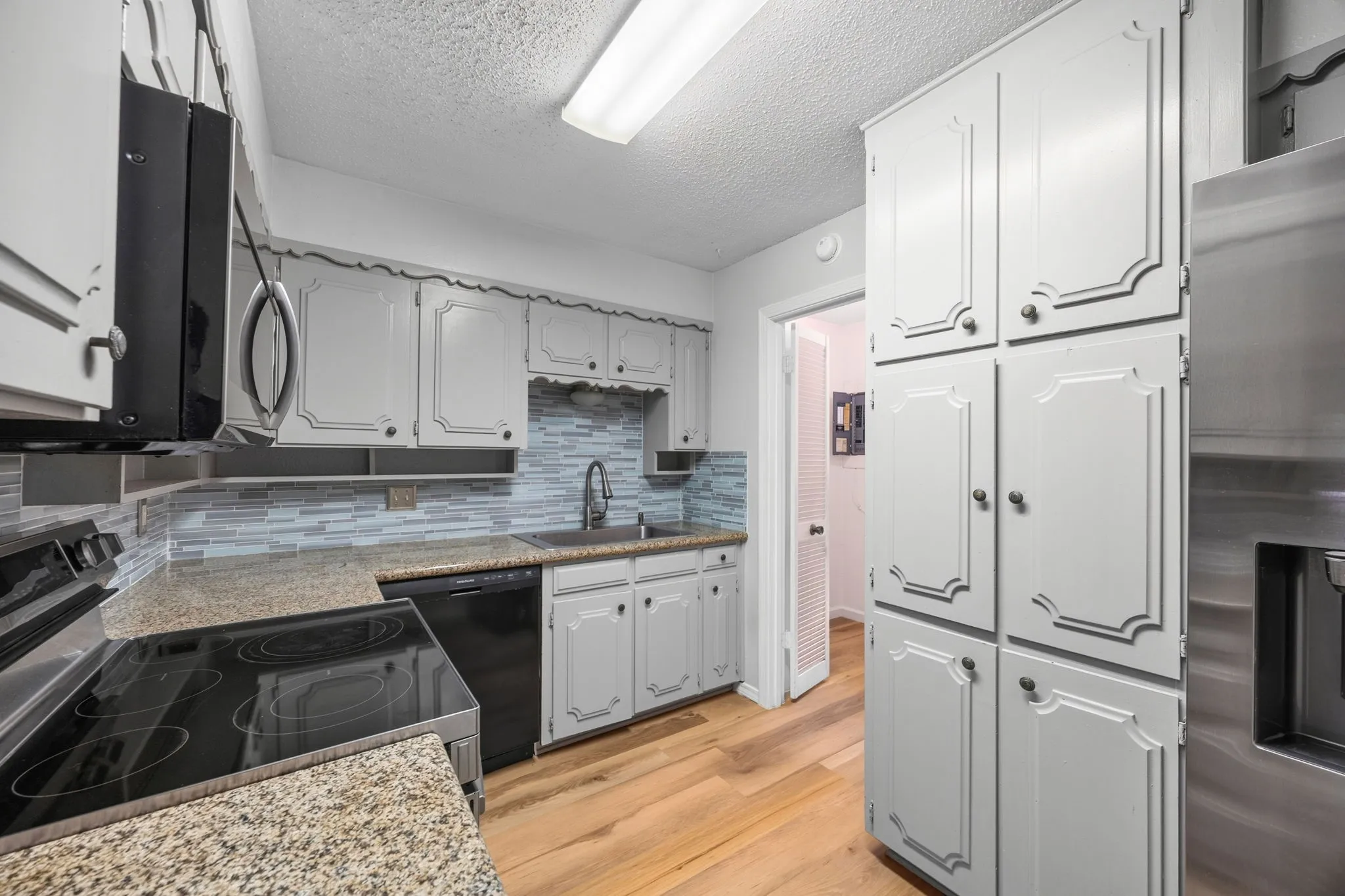 Kitchen featuring wood-finish flooring, gray perimeter cabinetry, granite-style countertops, a mosaic tile backsplash, and stainless steel appliances