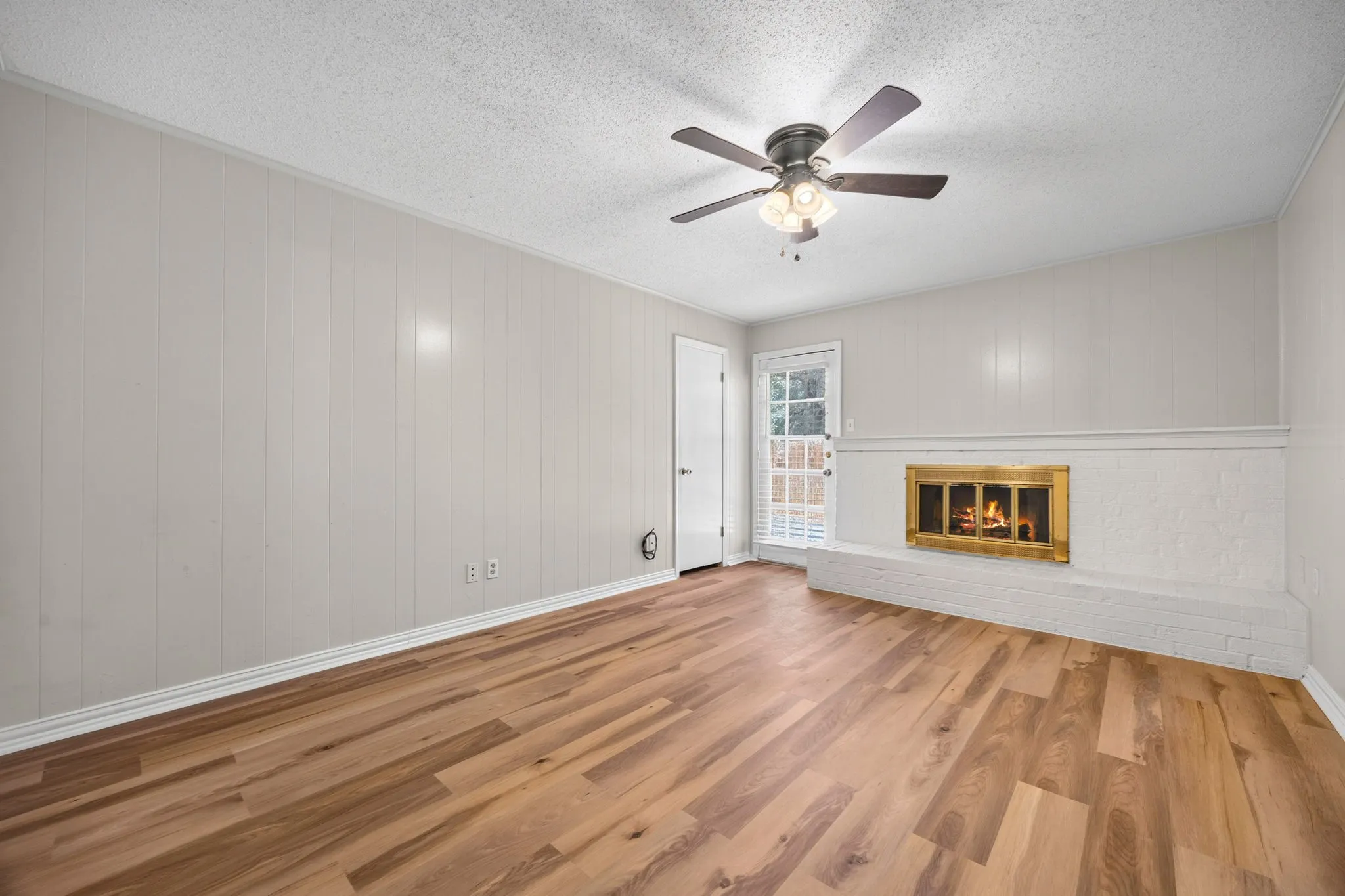 Living area featuring wood-finish flooring, a white painted brick fireplace with a brass firebox, and a ceiling fan with integrated lighting