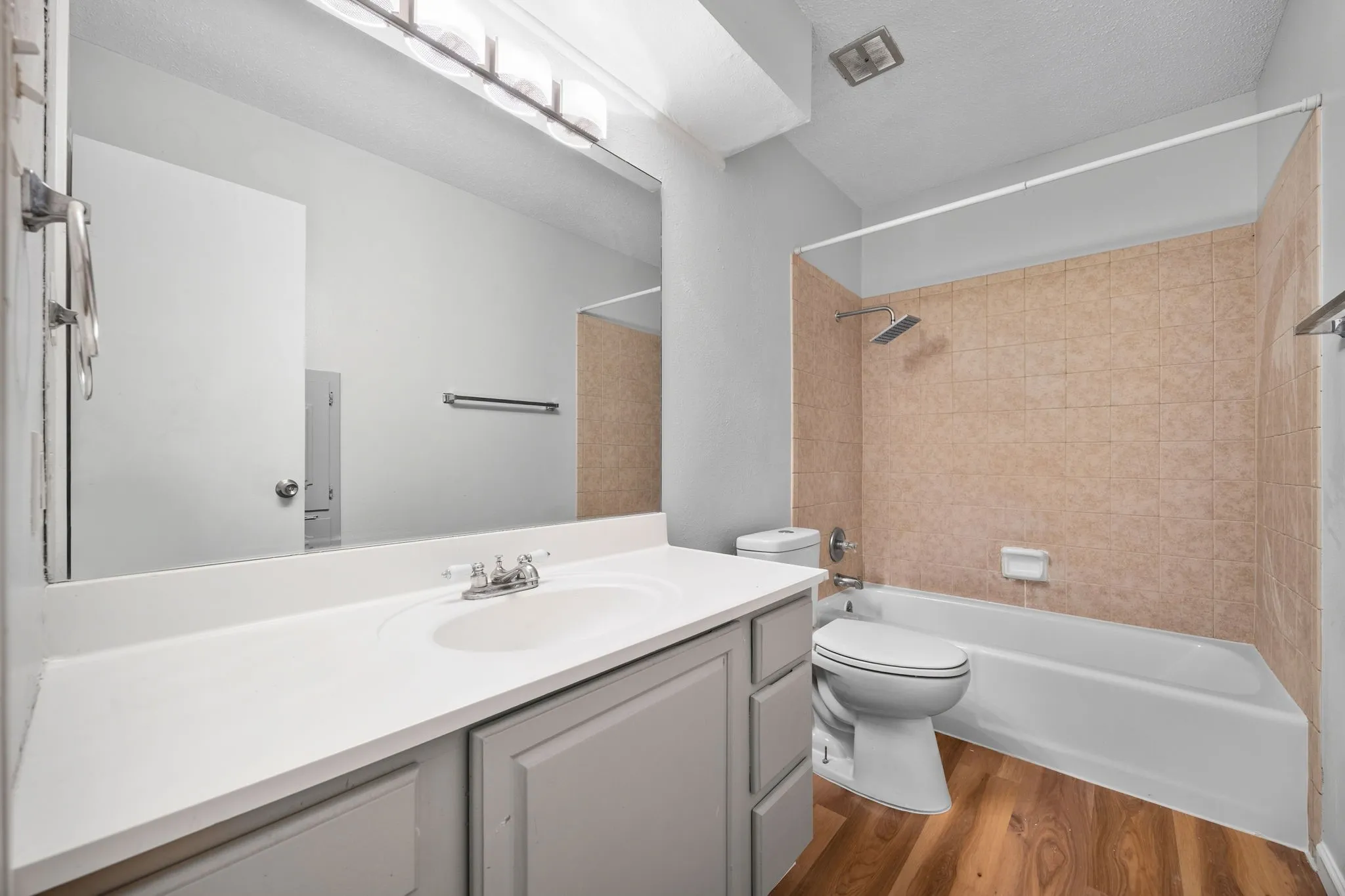 Bathroom featuring a single vanity with an integrated sink, a large wall-mounted mirror, a bathtub with a tiled surround, and wood-finish flooring