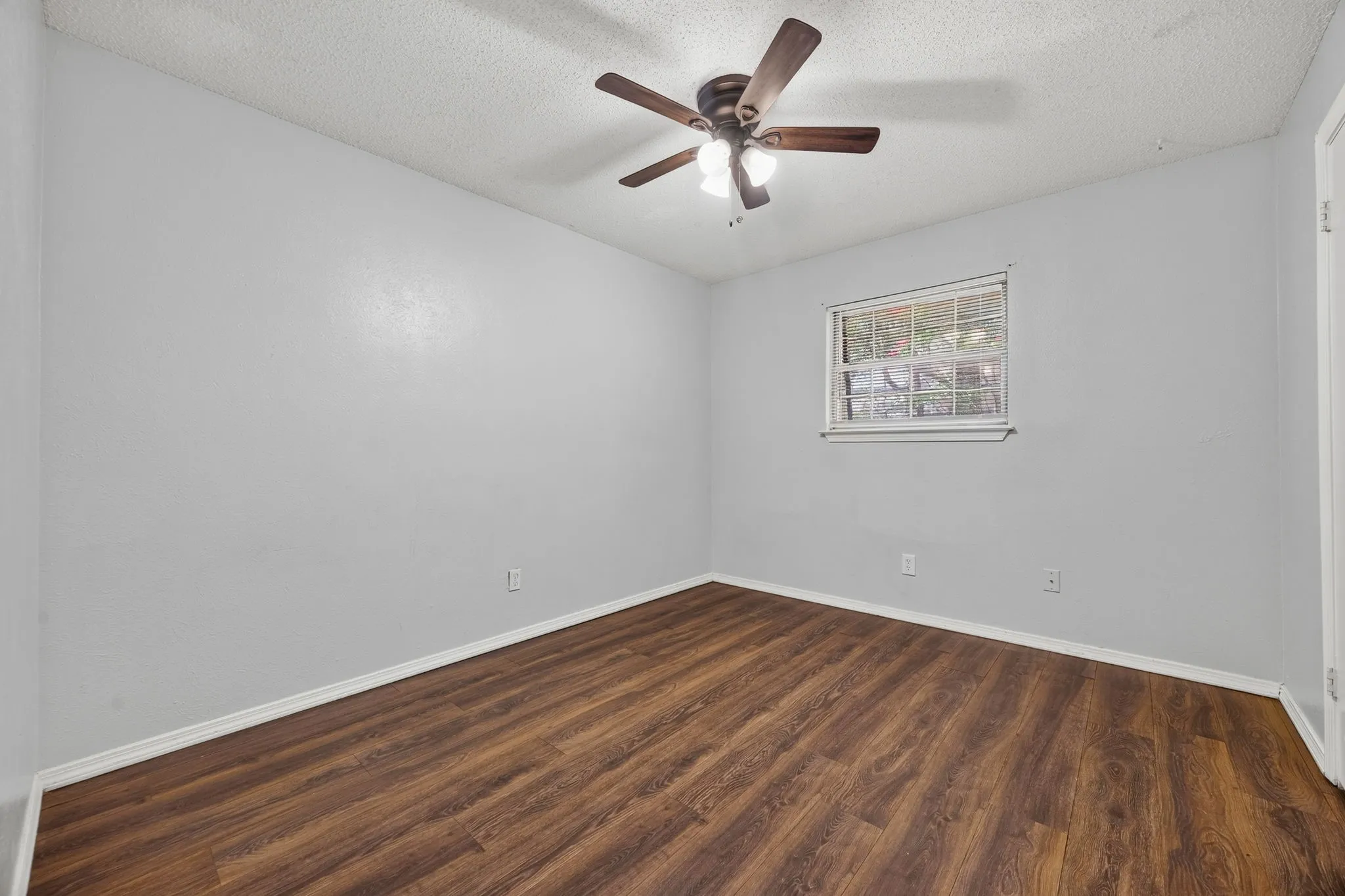Spacious bedroom showcasing warm wood-finish flooring, fresh white walls, ceiling fan with integrated lighting, and a fixed window with decorative grilles
