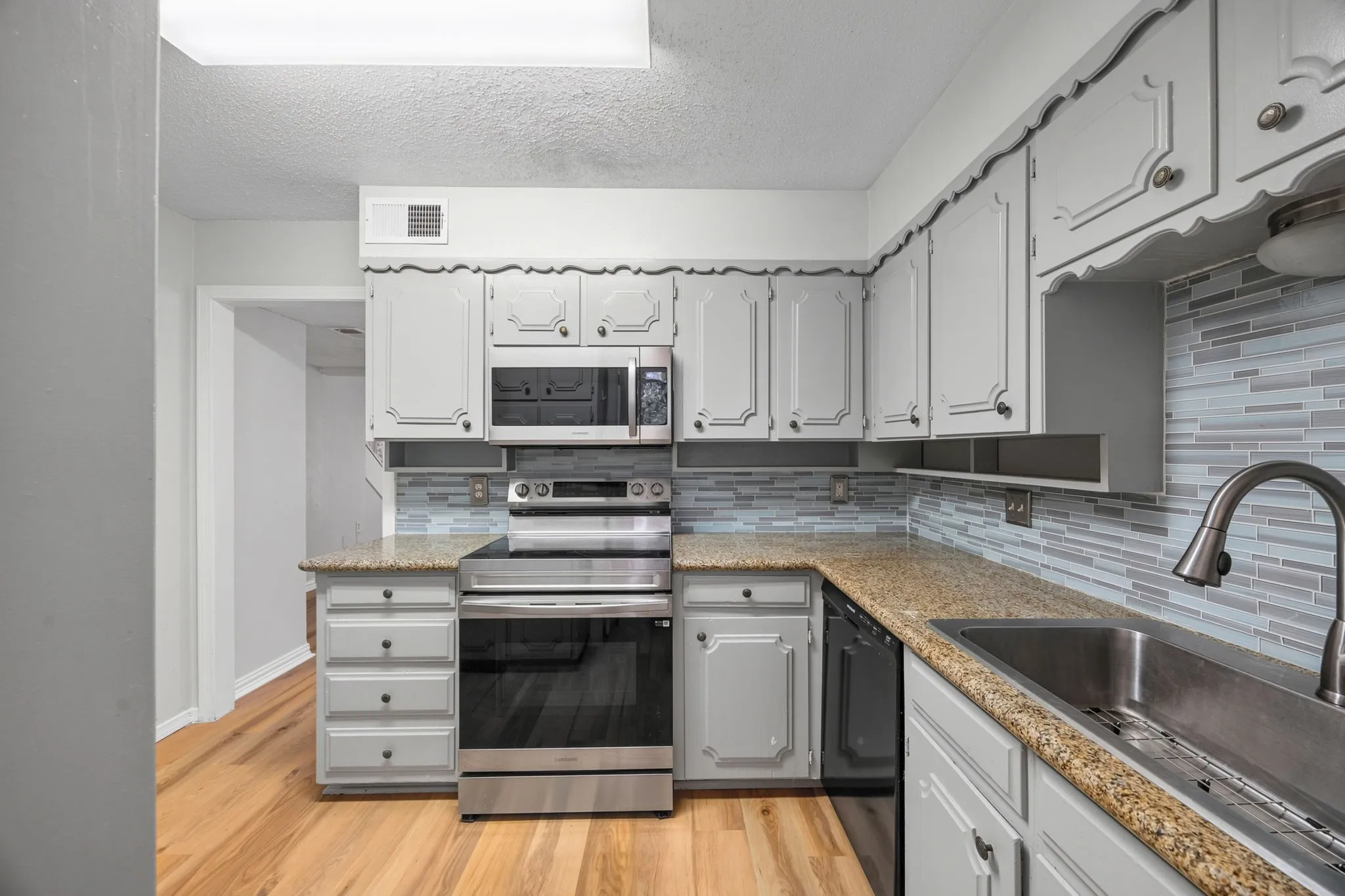 Kitchen featuring light gray cabinetry with decorative trim, granite countertops, stainless steel appliances, a horizontal tile backsplash, and light wood-finish flooring