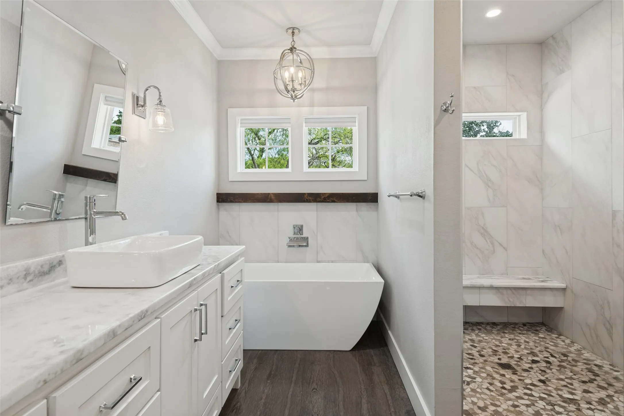 Freestanding soaking tub, dual windows with wood trim, wood-finish flooring, white vanity with stone countertop, and a walk-in shower with pebble tile floor and built-in bench