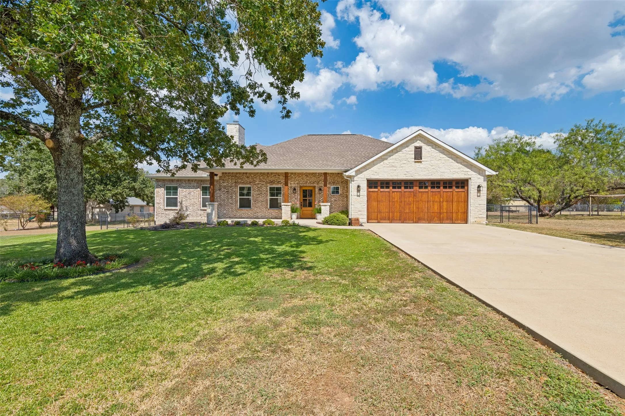 Brick exterior with natural wood garage doors and front entry, covered porch, and mature landscaping