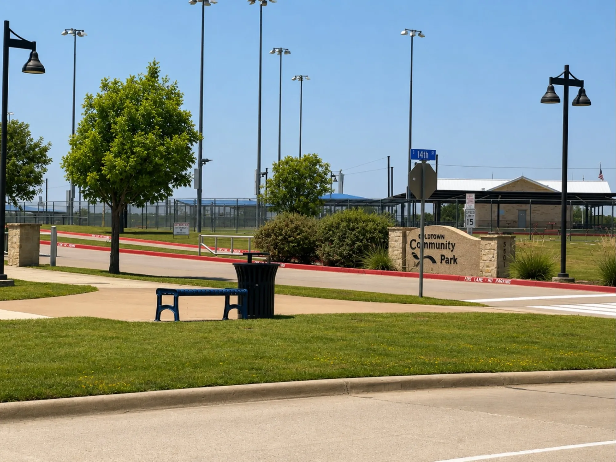 Community park entrance featuring a stone monument sign, paved pathways, mature landscaping, and overhead lighting fixtures