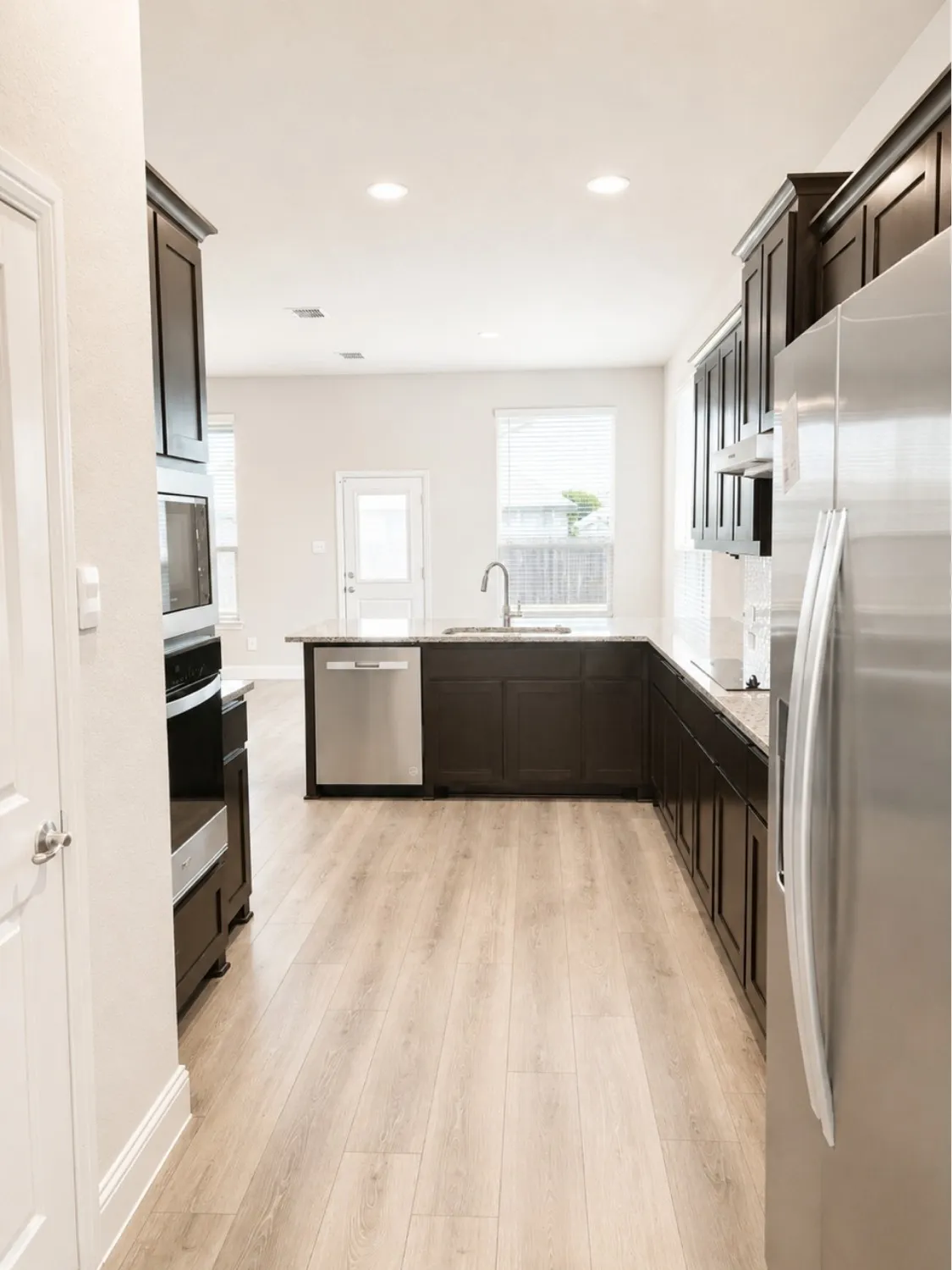 Modern galley kitchen featuring dark cabinetry with integrated hardware, stainless steel appliances including a side-by-side refrigerator and built-in microwave, a contrasting dark island with undermount sink and prep space, recessed ceiling lighting, and light wood-finish flooring throughout