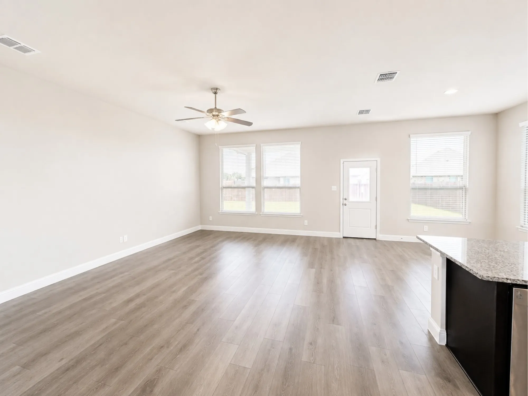 Spacious living area featuring light wood-finish flooring, a ceiling fan with integrated lighting, multiple windows with blinds, a white door providing exterior access, and a kitchen island with a granite countertop