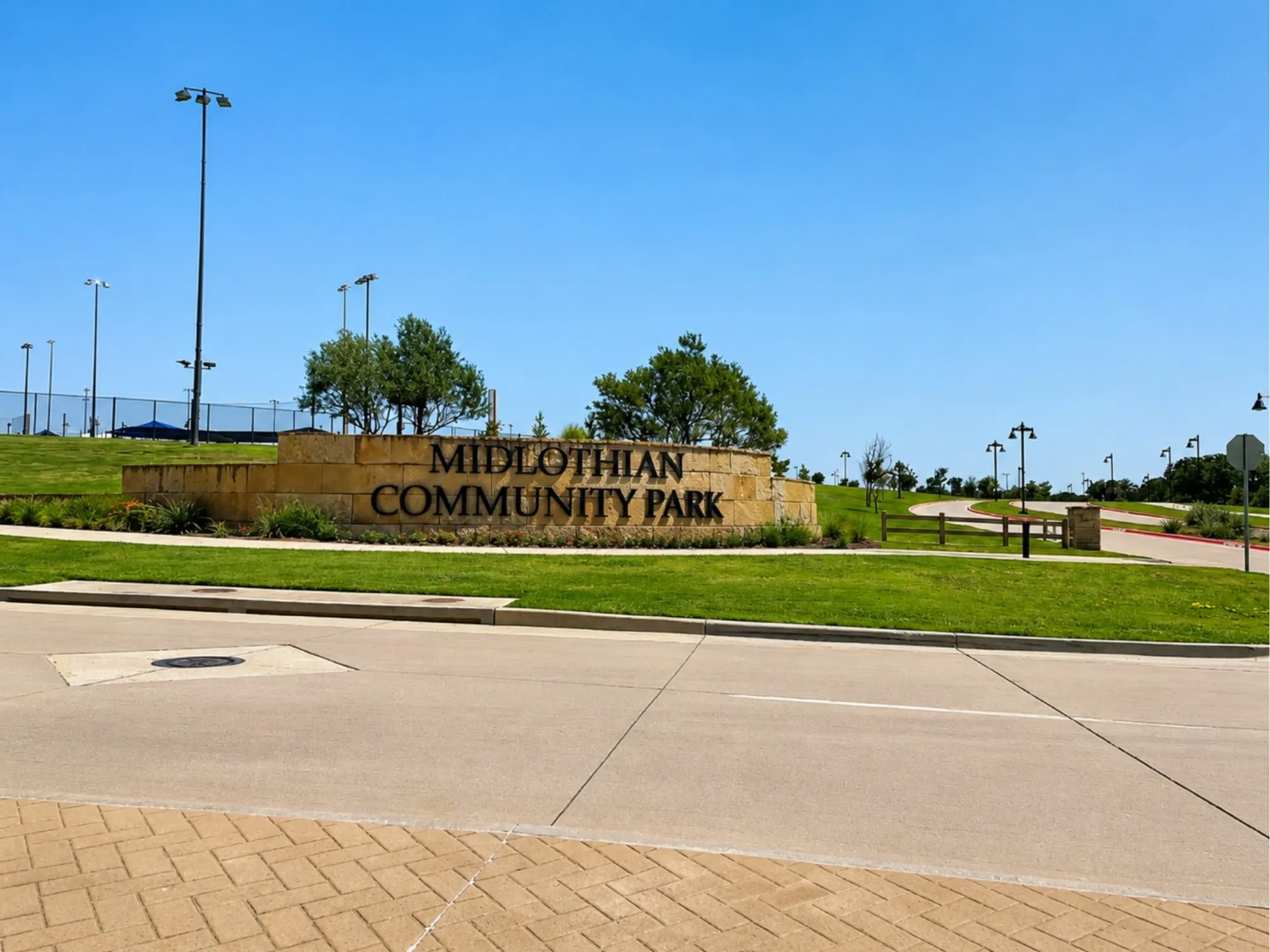 Midlothian Community Park entrance featuring a welcoming stone monument sign, expansive manicured lawn, mature landscaping with established trees, professional lighting infrastructure, and well-defined paved pathways throughout the grounds