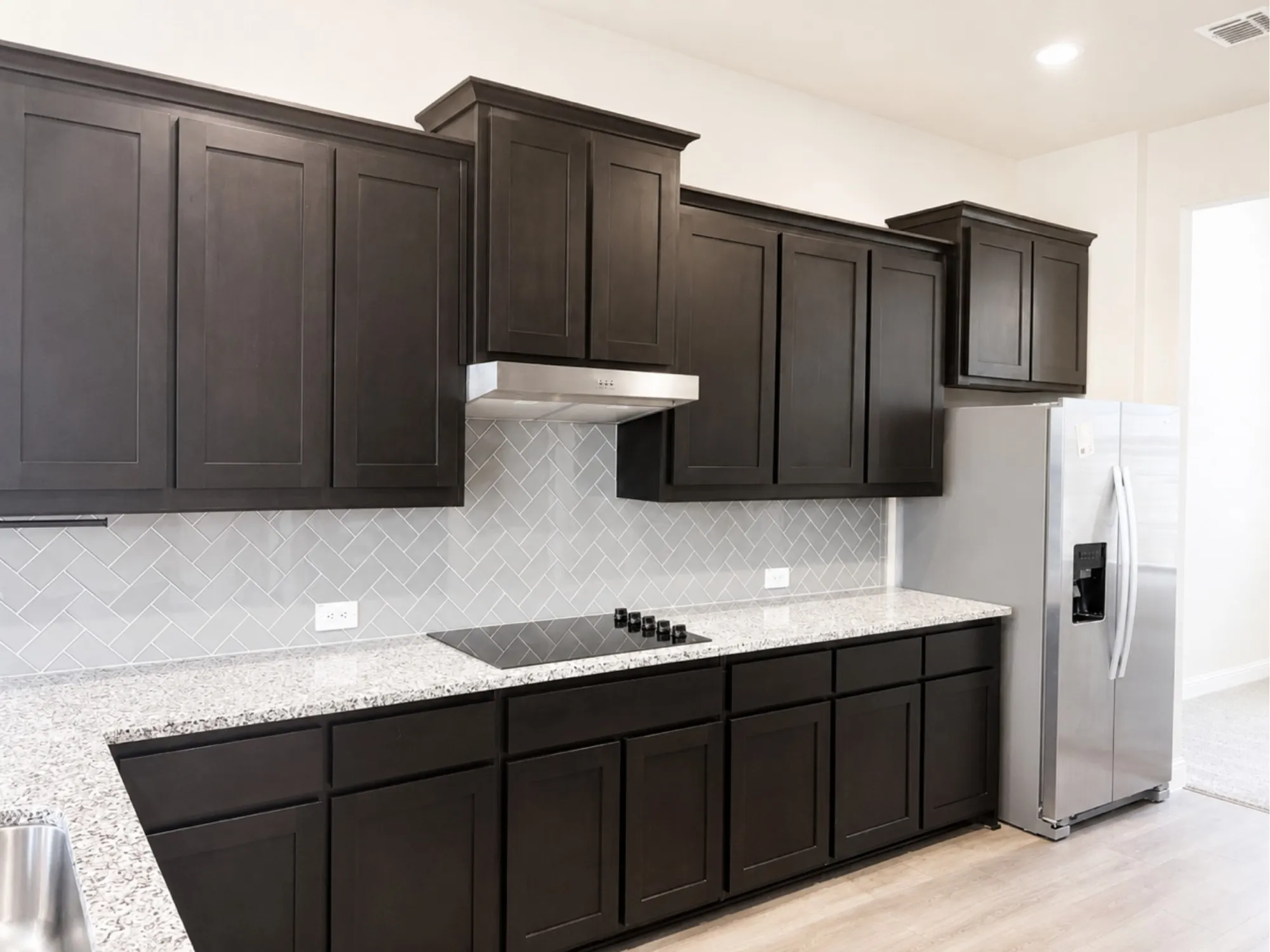 Modern kitchen with dark wood-finish cabinetry, light speckled countertops, and a herringbone tile backsplash