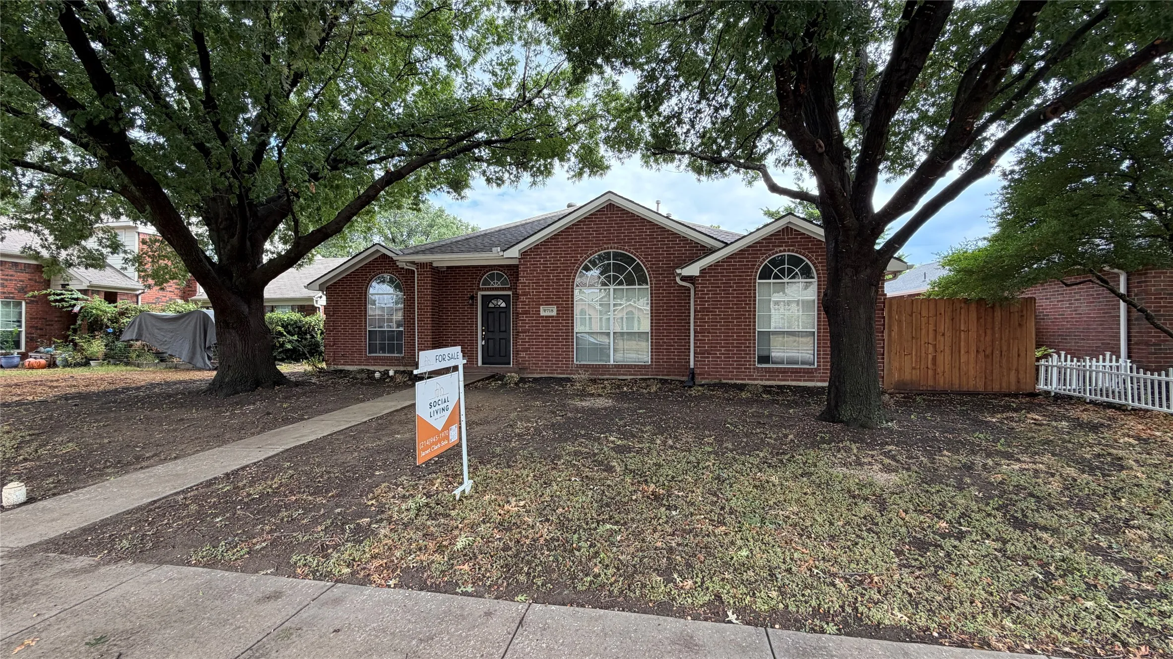 Brick exterior with arched windows, mature shade trees, a concrete walkway, and a partial wood fence