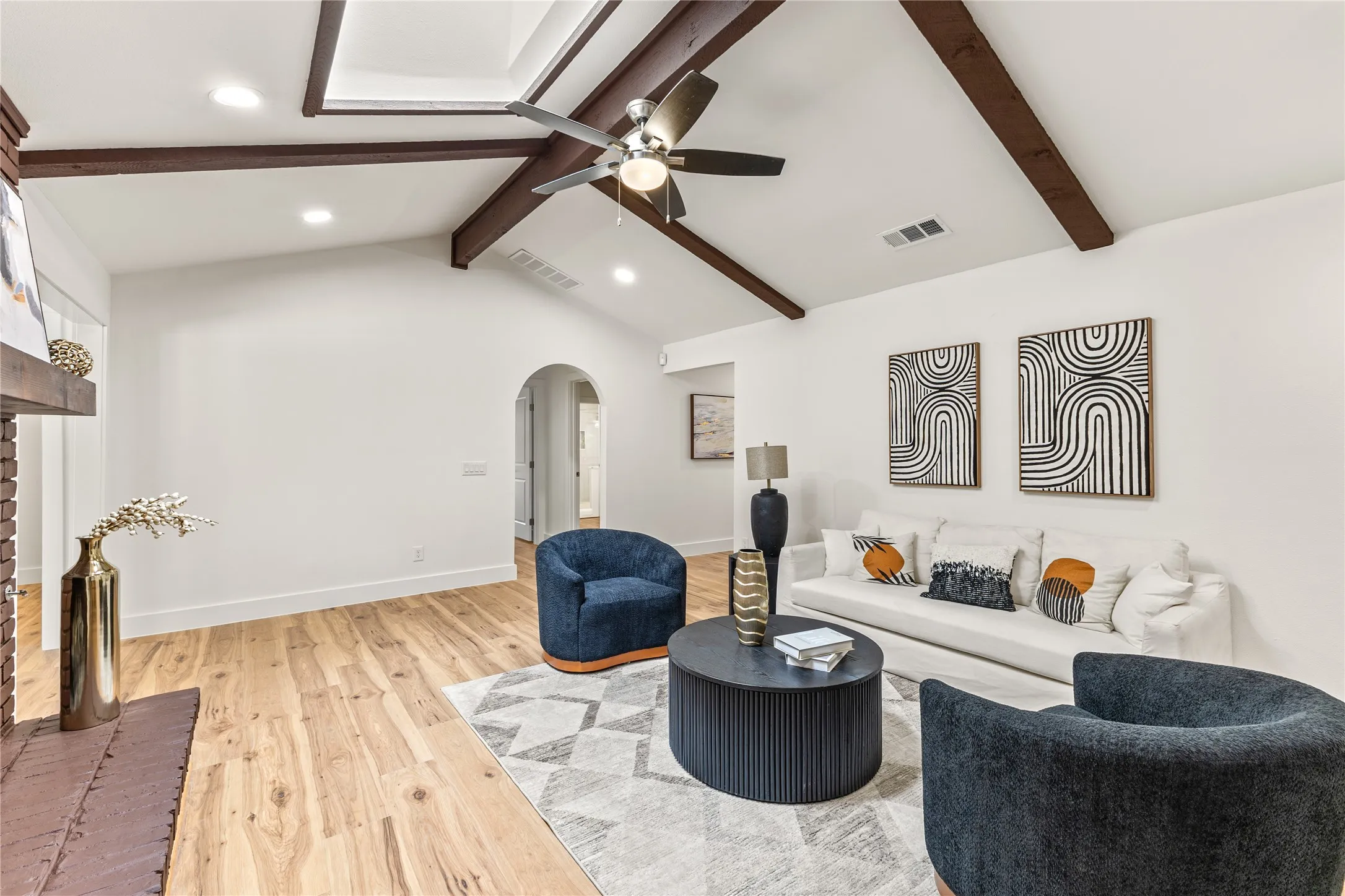 Vaulted ceiling with exposed wood beams, wood-finish flooring, brick fireplace, recessed lighting, and an arched doorway