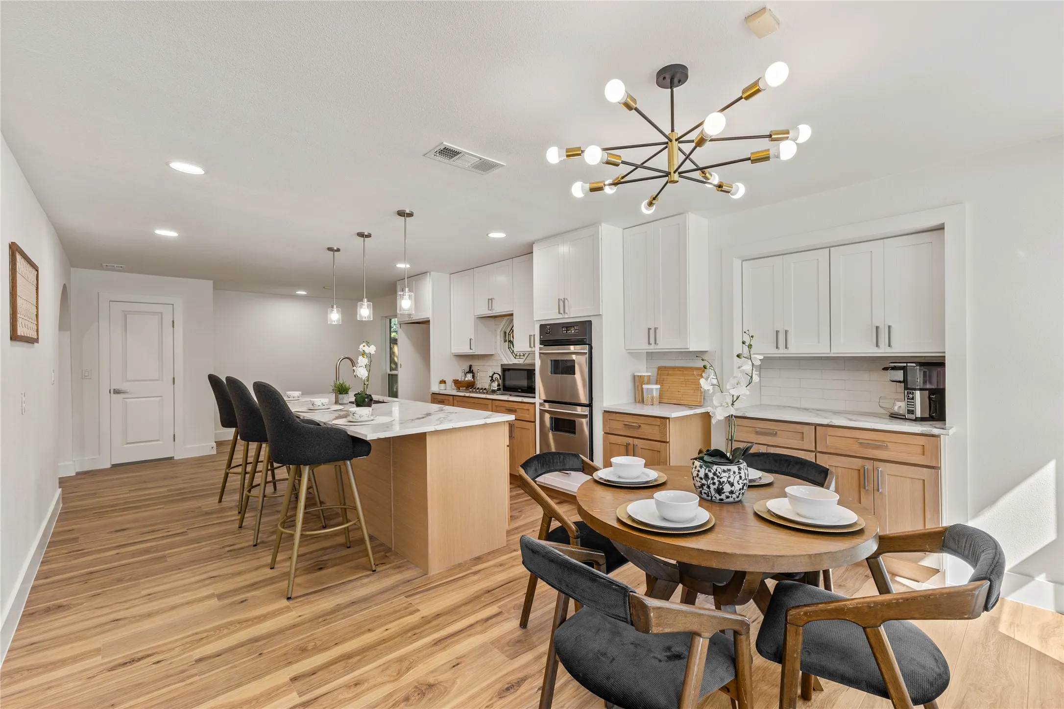 Modern kitchen featuring wood-finish flooring, a kitchen island with pendant lighting, white upper cabinetry, light wood lower cabinetry, and stainless steel double ovens
