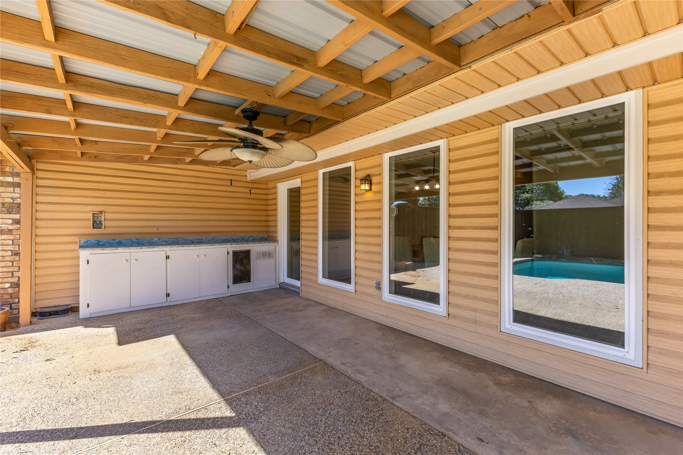 Covered patio with exposed wood beam ceiling, ceiling fan with integrated lighting, built-in cabinetry with counter space, and three large windows