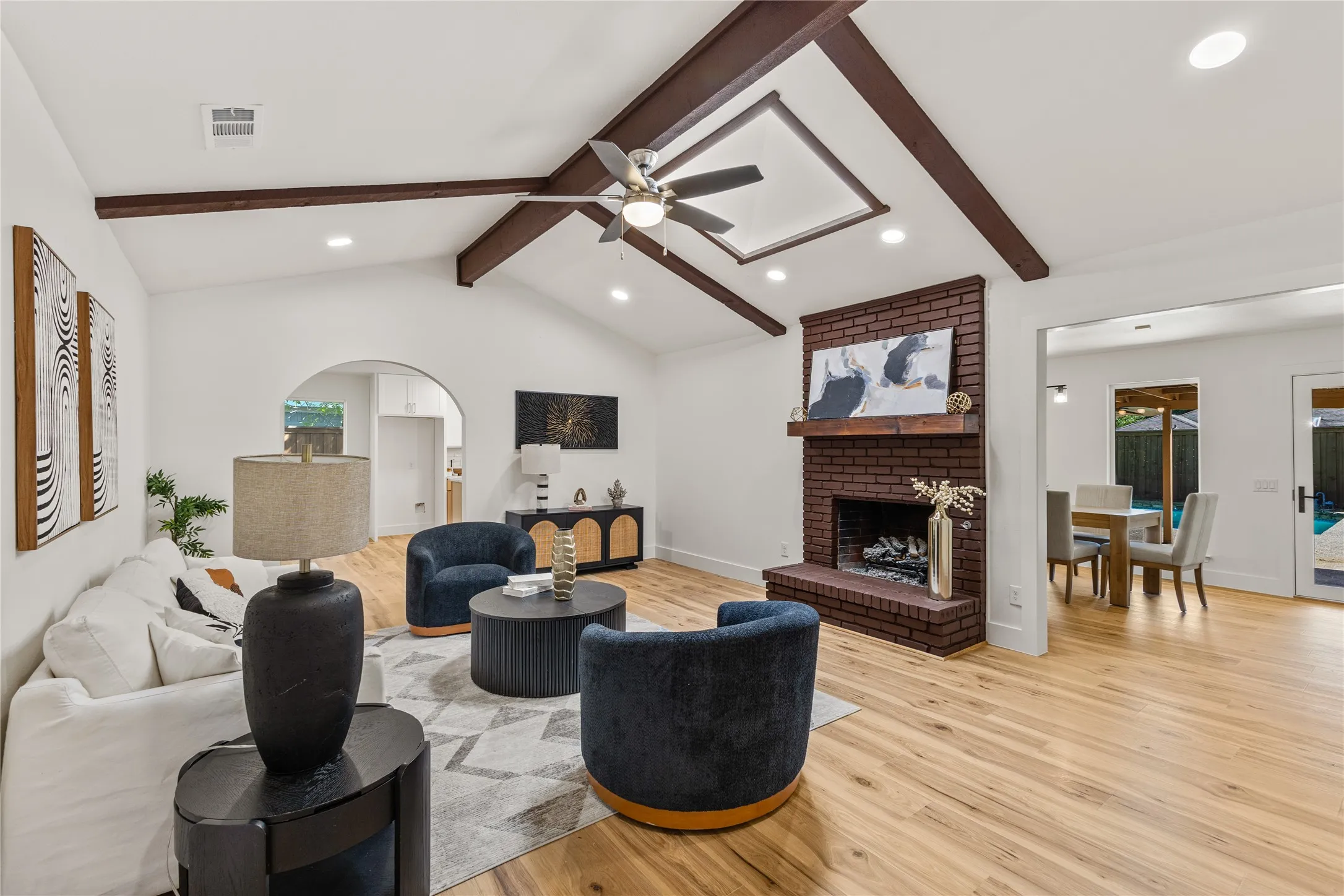 Vaulted ceiling with exposed wood beams, brick-clad fireplace with mantel, recessed lighting, wood-finish flooring, and an arched doorway