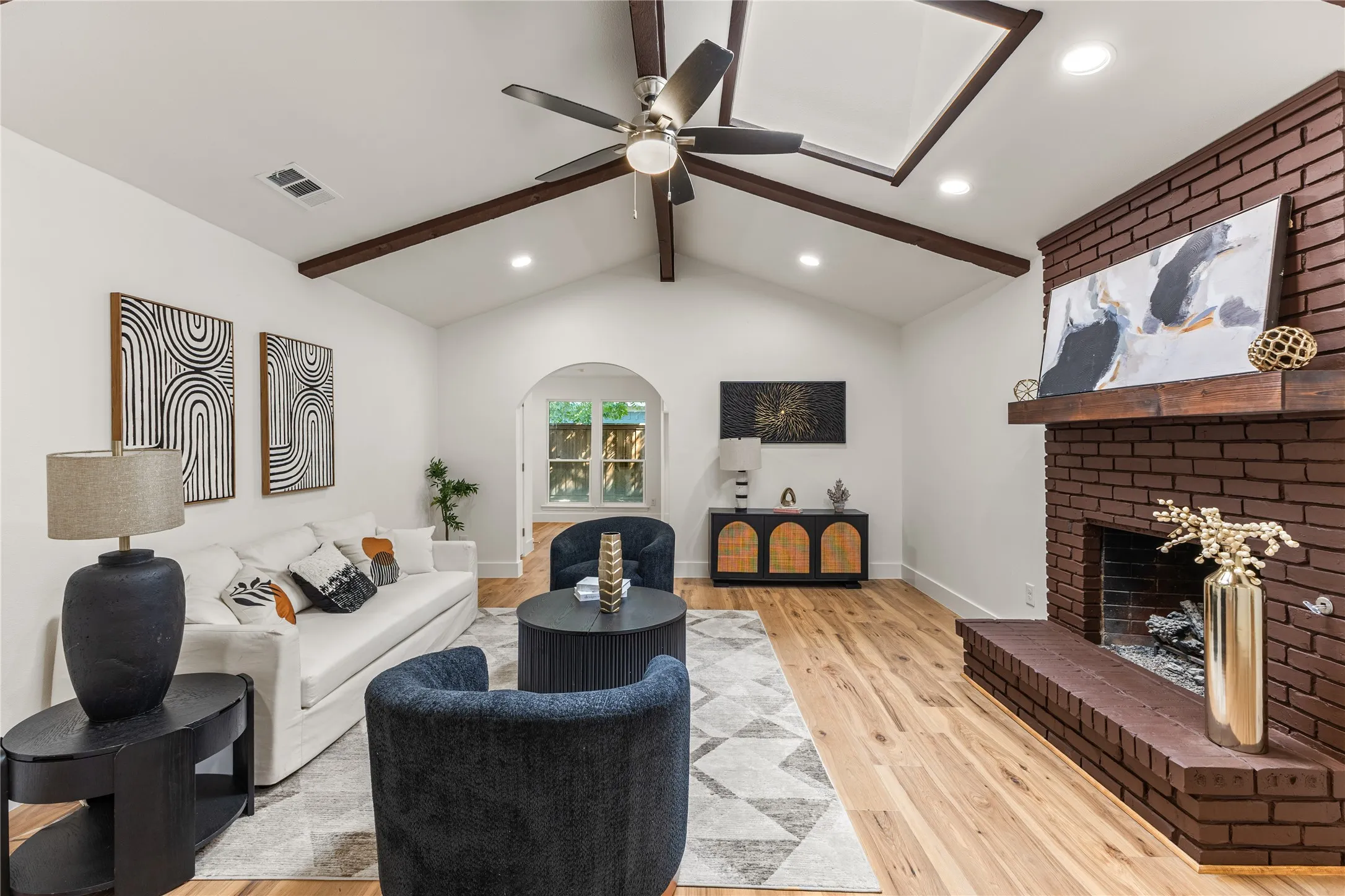 Vaulted ceiling with exposed wood beams, a prominent brick fireplace, wood-finish flooring, and recessed lighting