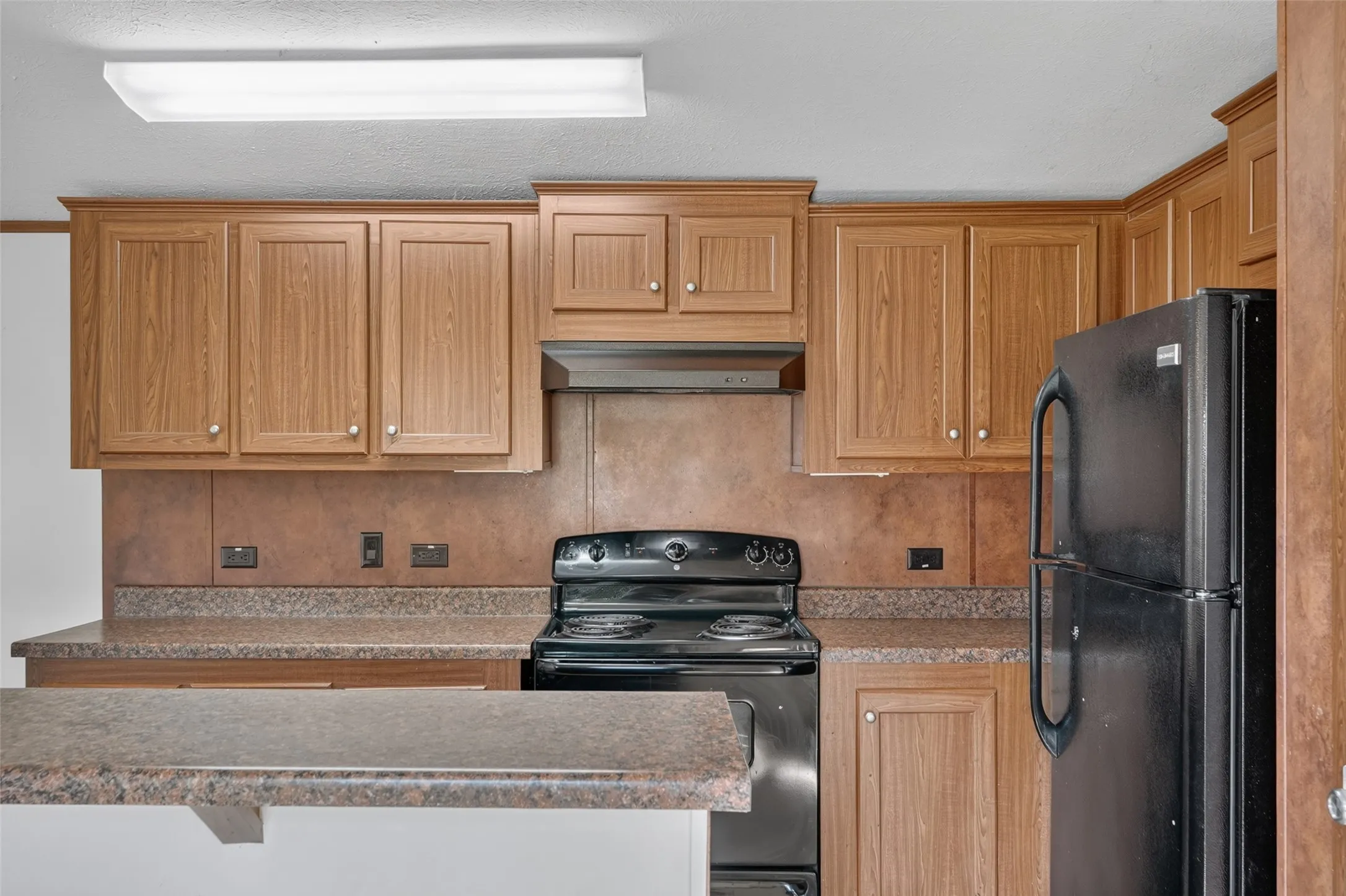 Kitchen featuring light wood-finish cabinetry, black electric range, black refrigerator, and an extended countertop