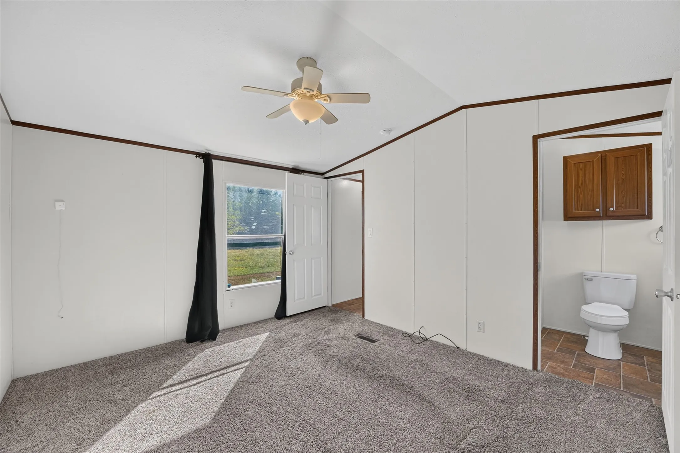 Carpet flooring, ceiling fan with light fixture, window with outdoor scenery, and an attached bathroom with tile flooring and wood-finish cabinetry