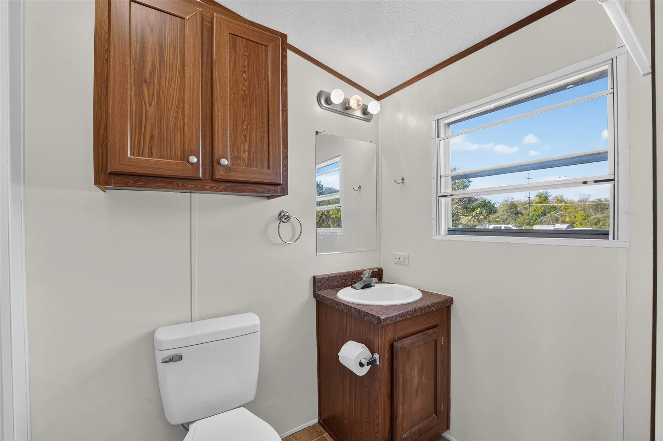 White toilet with a wall-mounted cabinet, wood-finish vanity, and a window