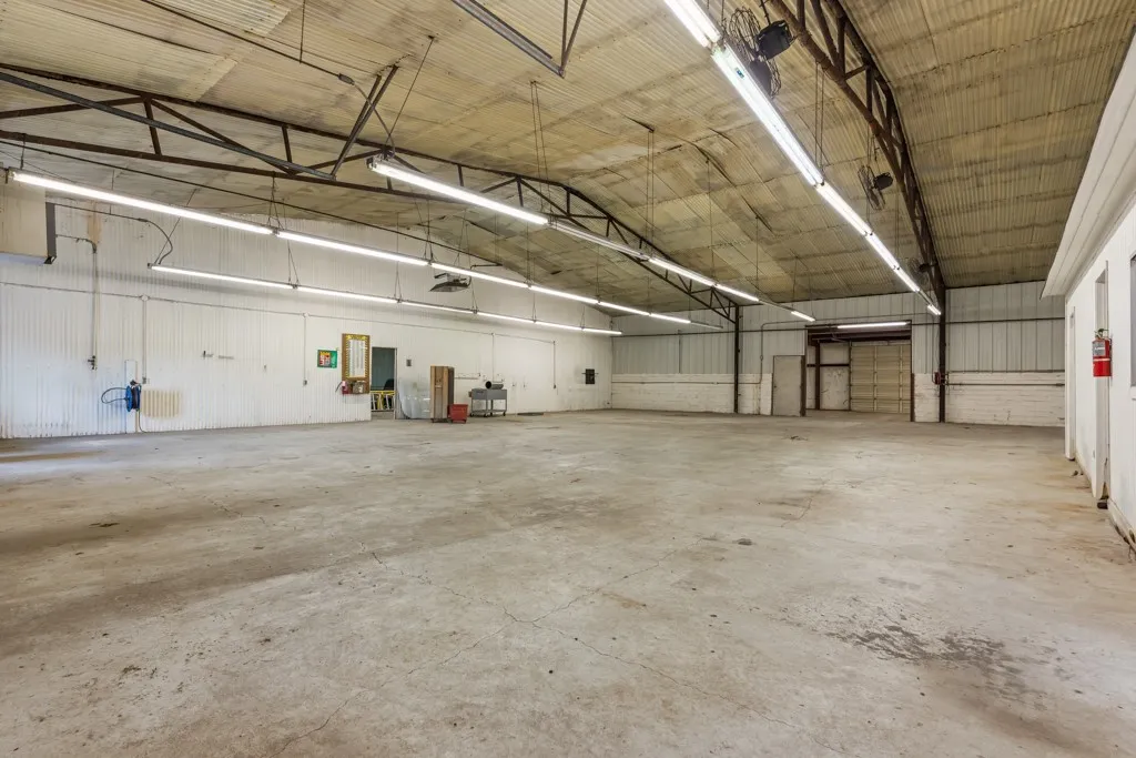 Expansive open warehouse space featuring concrete flooring, corrugated metal walls, exposed structural beams, and fluorescent overhead lighting
