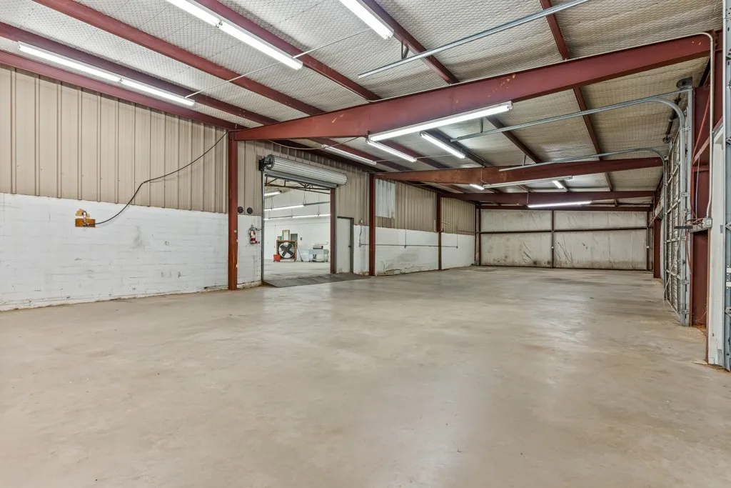 Spacious warehouse featuring a concrete floor, exposed structural beams, corrugated metal walls, overhead fluorescent lighting, and two roll-up doors