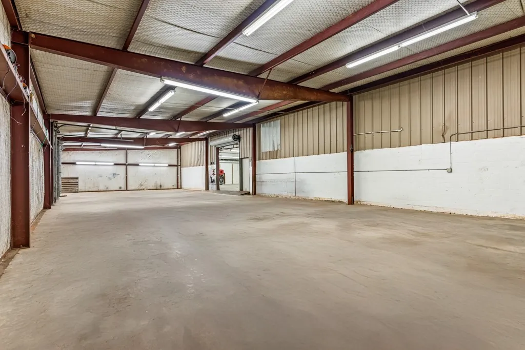 Spacious interior featuring exposed structural beams, a concrete floor, corrugated metal walls, and fluorescent lighting fixtures