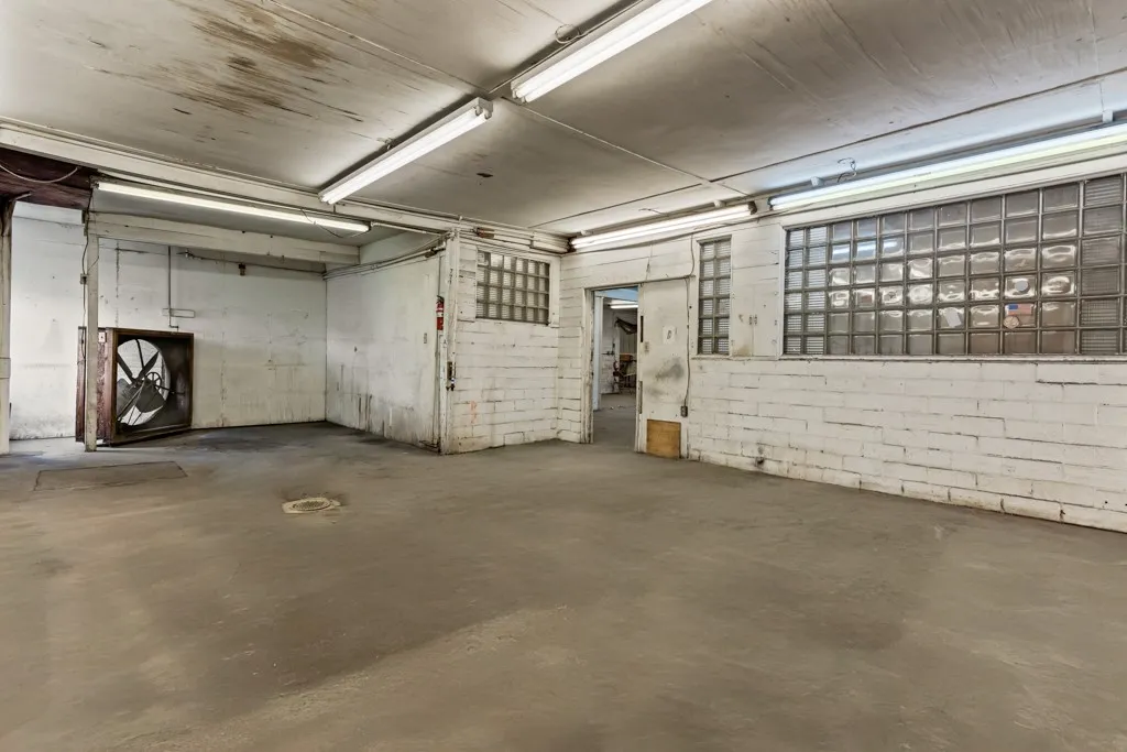 Concrete flooring, exposed ceiling with fluorescent lighting, painted brick walls, glass block windows, and an industrial-style wall fan