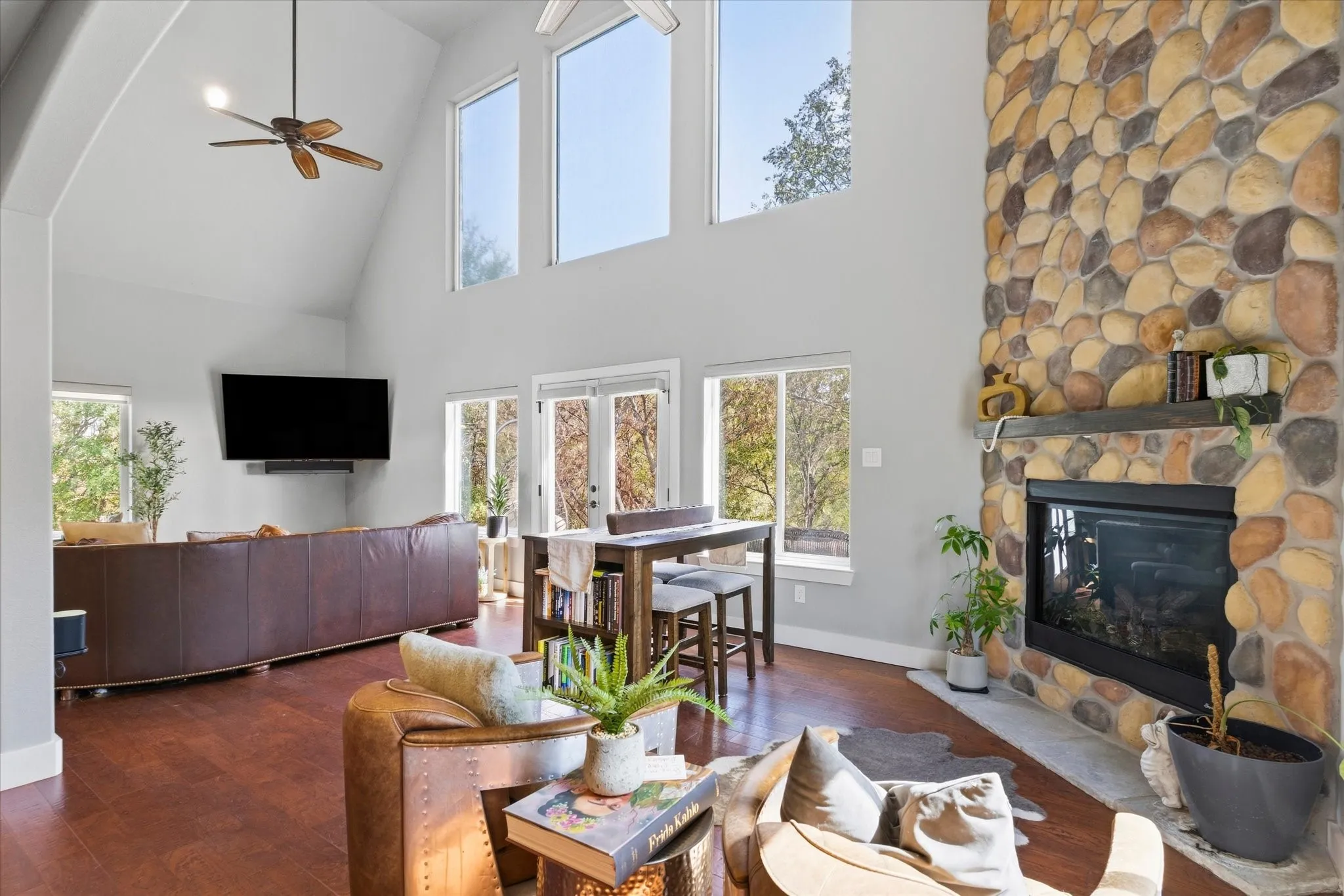 Vaulted ceiling with ceiling fan, large stone fireplace with mantel, dark wood-finish flooring, and multiple windows