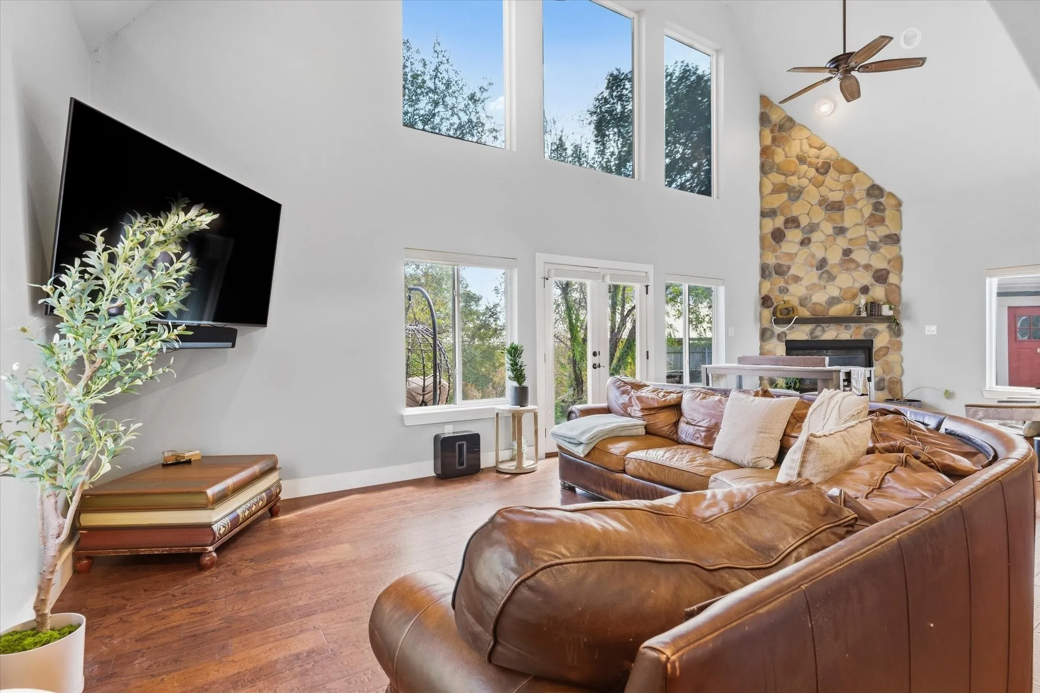 Vaulted ceiling with a ceiling fan, stone fireplace with a mantel, multiple large windows, and a glass-paneled door
