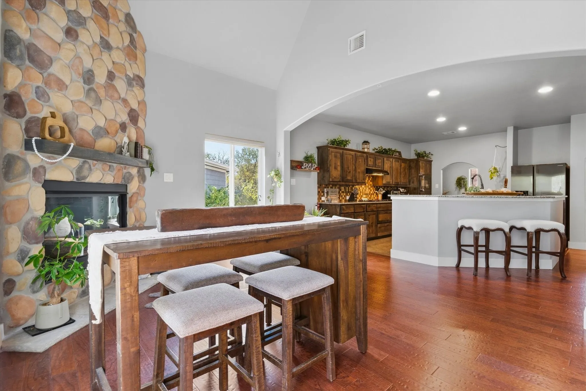 Stone-faced fireplace with a substantial mantel, vaulted ceiling, wood-finish flooring, and an open archway to the kitchen