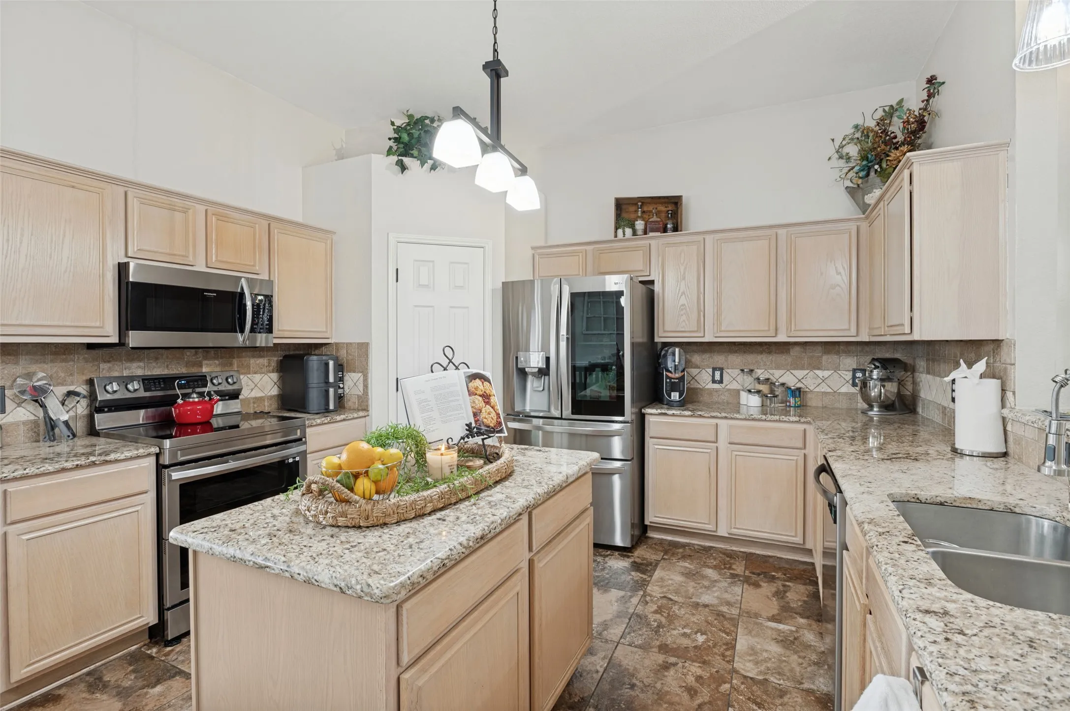 Kitchen featuring a central island, light wood-finish cabinetry, stainless steel appliances, speckled countertops, and a tiled backsplash