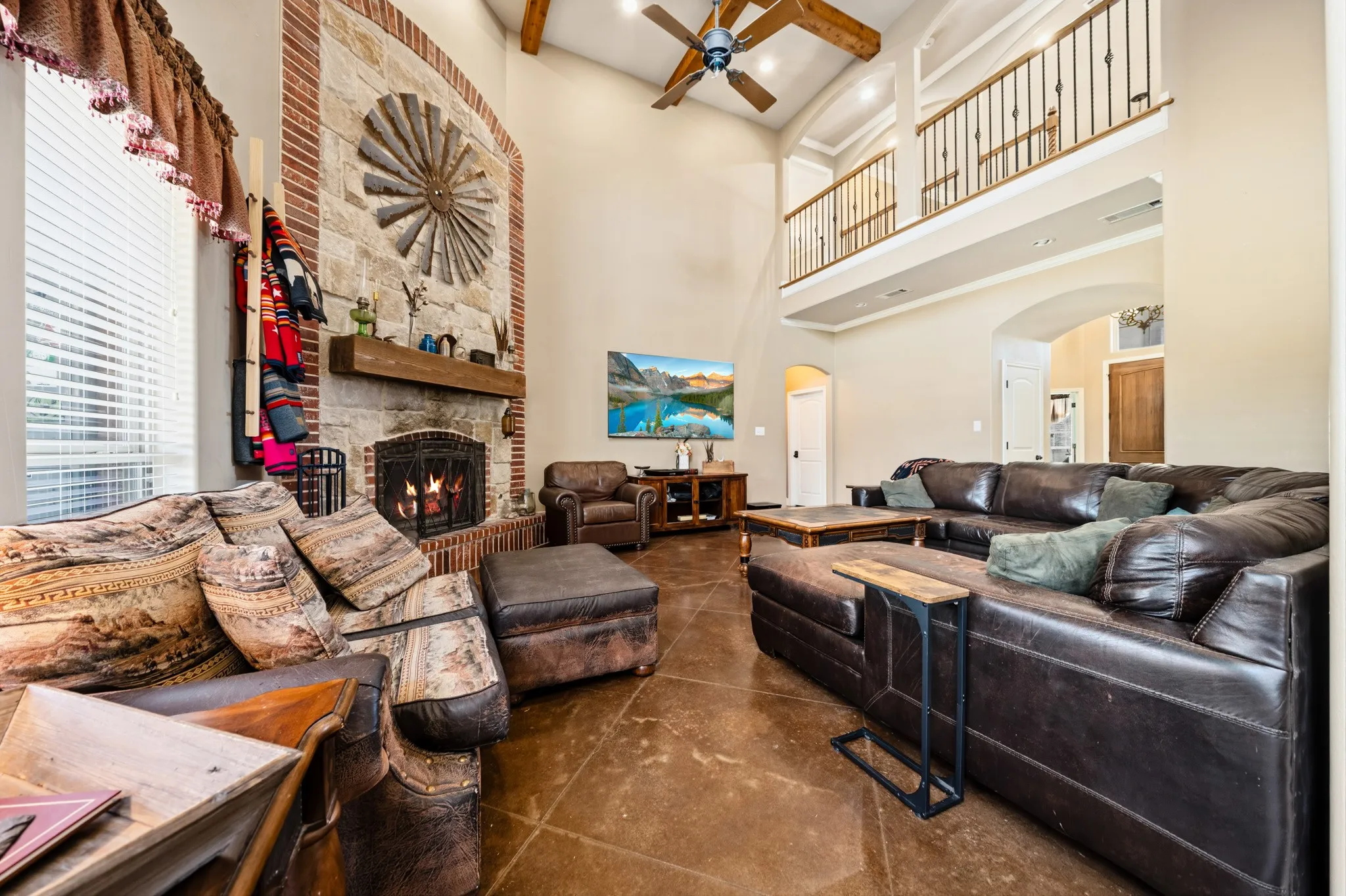 Vaulted ceiling with exposed wood beams, stone fireplace with a wood mantle, upper-level balcony with metal spindle railing, and stained concrete flooring