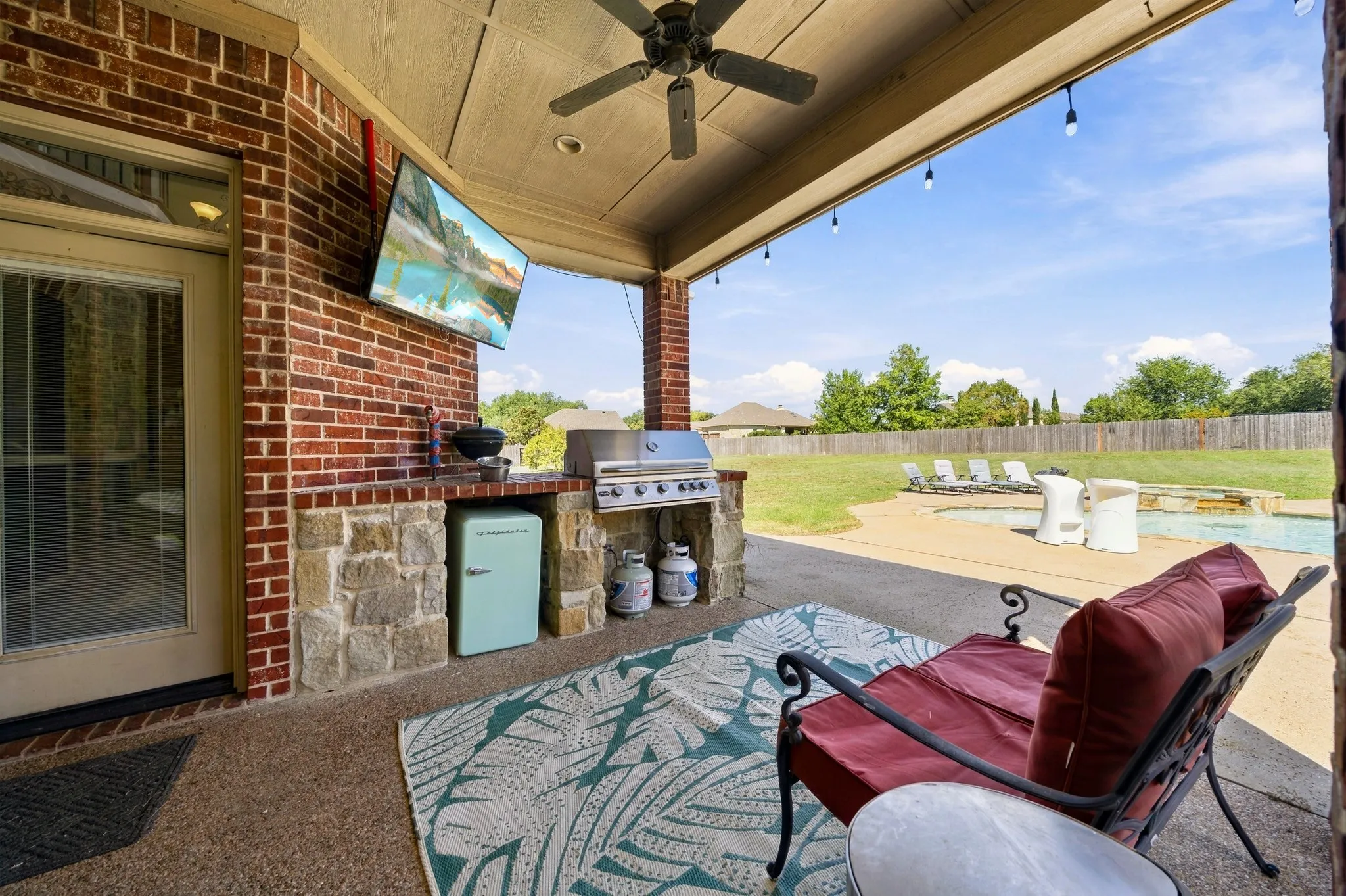 Covered patio featuring a built-in outdoor kitchen, ceiling fan, and string lights, overlooking a backyard with a swimming pool