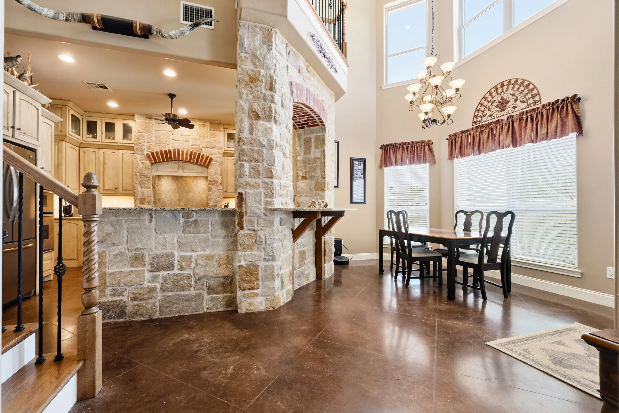 Two-story ceiling, stone accent walls and archway, wood-finish flooring, staircase with wrought iron balusters, and a kitchen featuring light-toned cabinetry