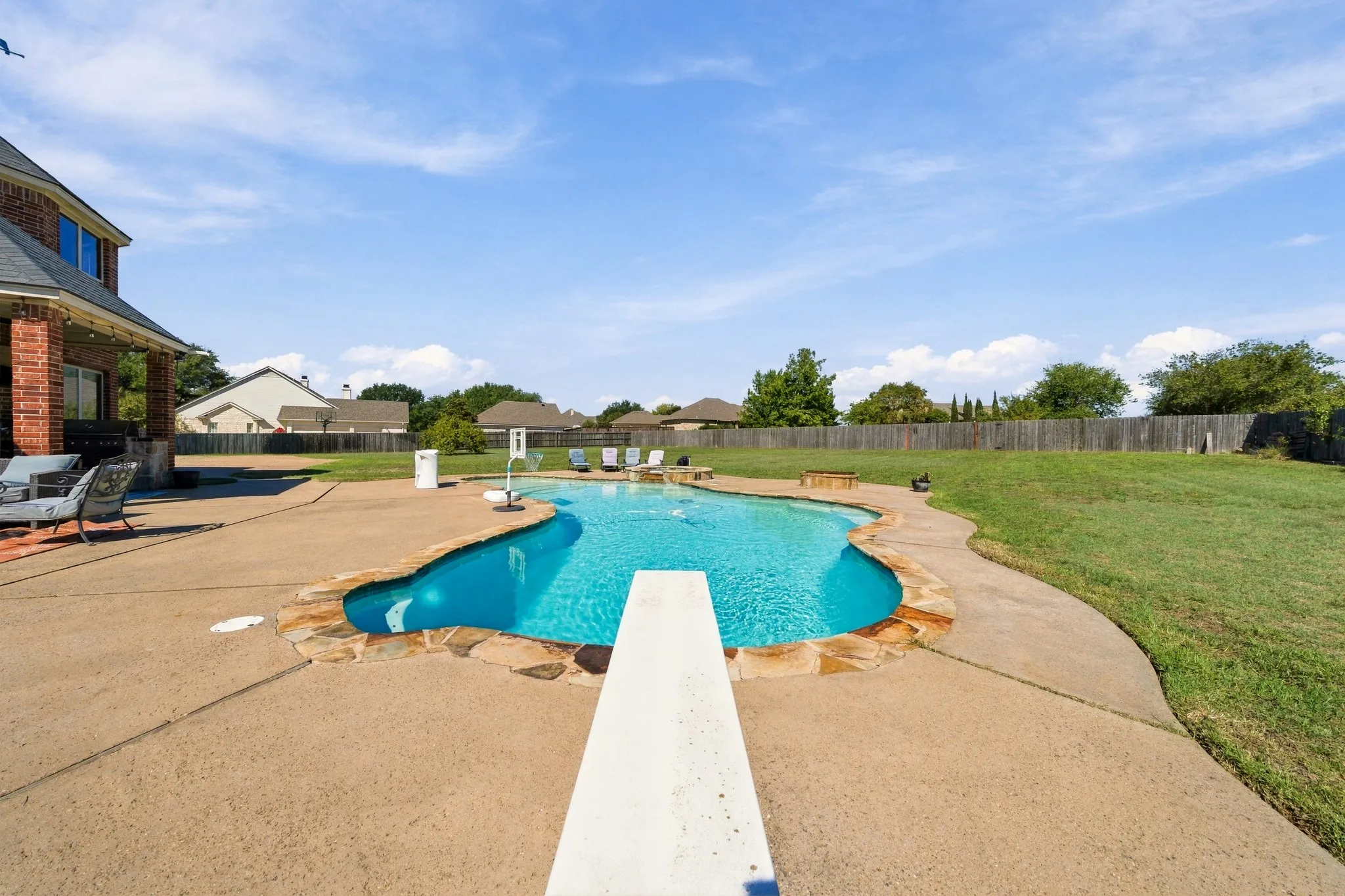 Curved swimming pool with stone coping, diving board, and expansive concrete patio