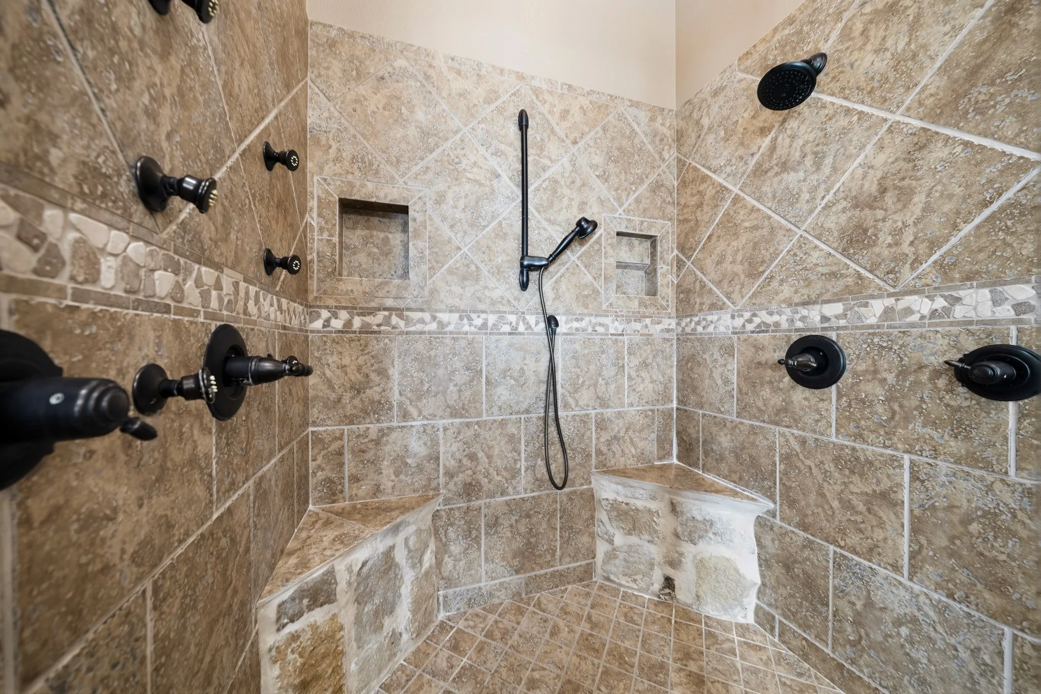 Tiled shower enclosure featuring multiple shower heads, a handheld shower, integrated tiled seating, and two recessed niches
