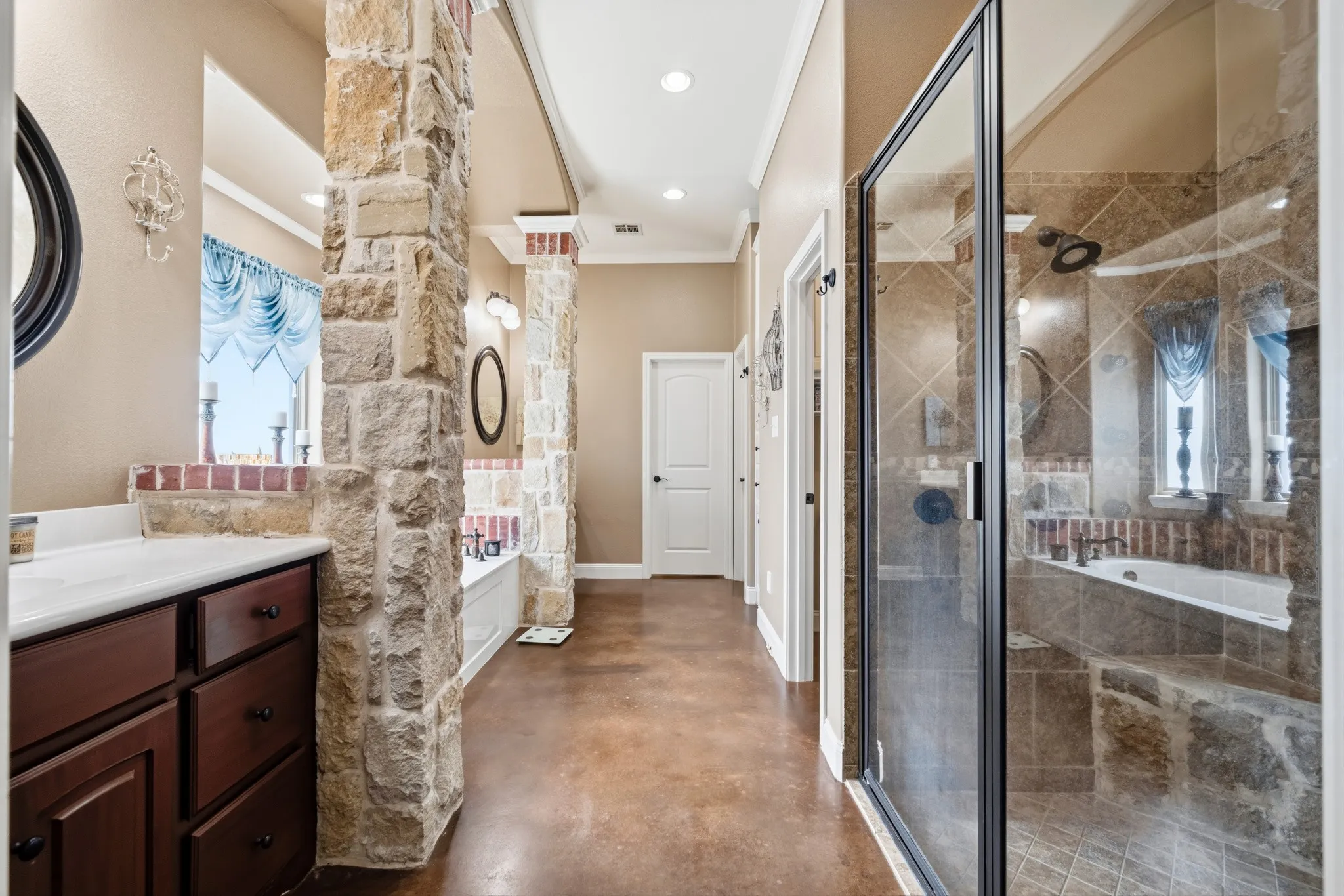 Walk-in shower with glass enclosure, soaking tub with tile surround, stone-clad architectural columns, dark wood-finish vanity with white countertop, and concrete-finish flooring