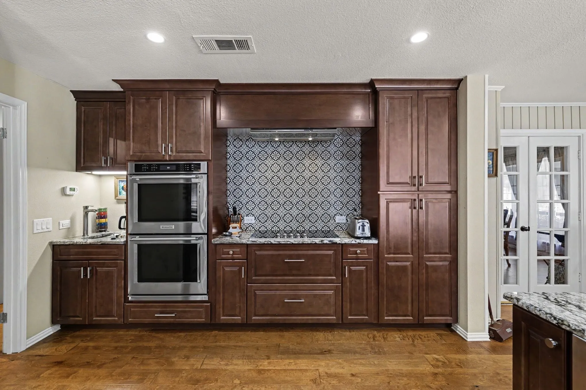 Wood-finish flooring, dark wood cabinetry, stainless steel double wall ovens, patterned tile backsplash, and light-toned countertops