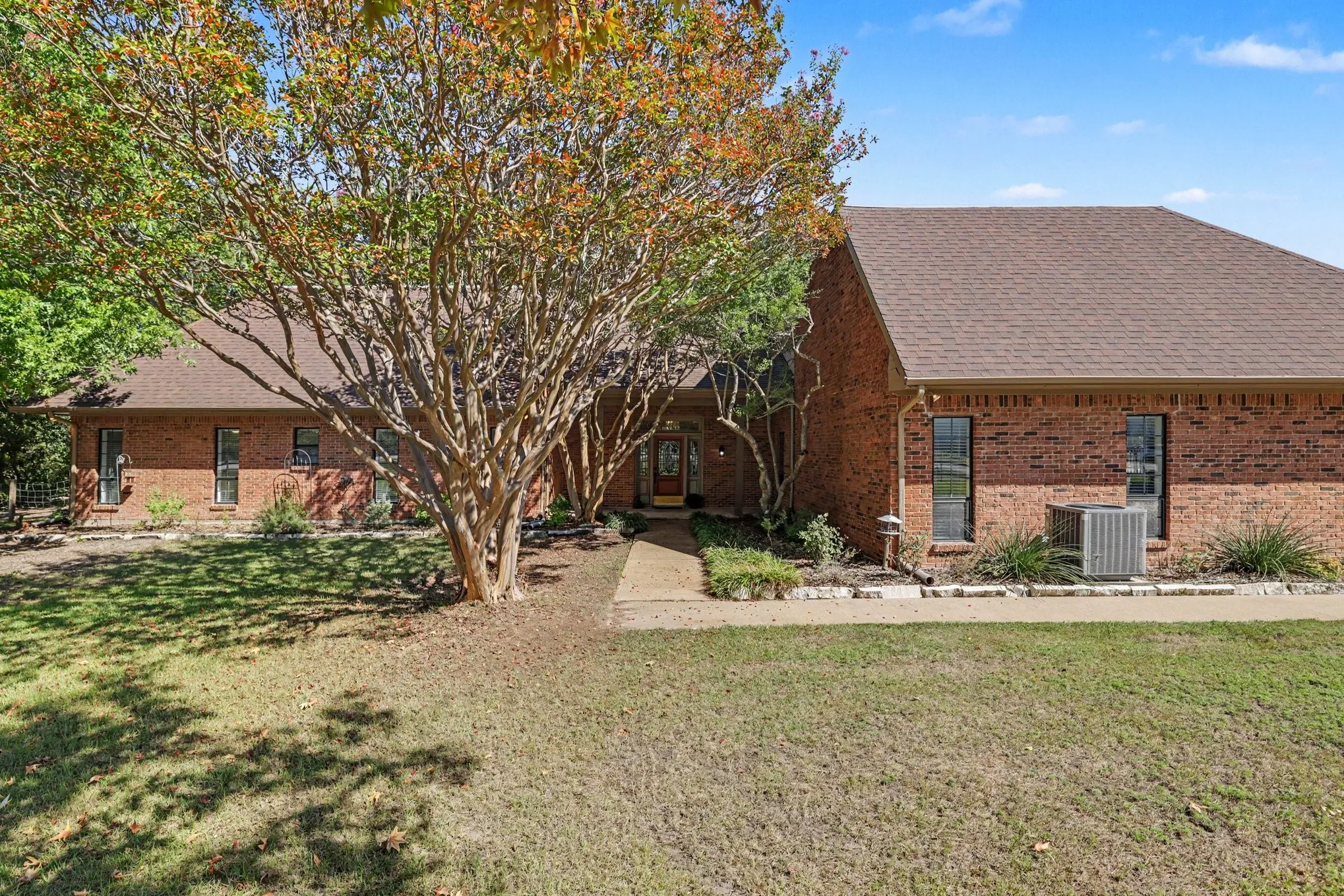 Brick exterior with a brown shingle roof, mature trees, and a concrete walkway