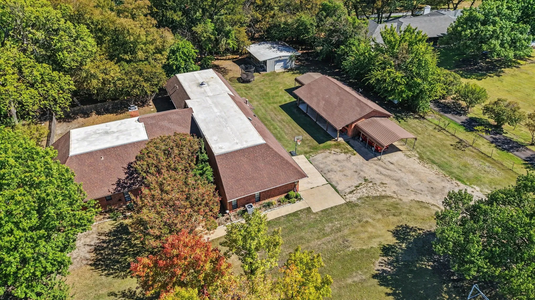 Expansive property featuring multiple structures, mature trees, a paved driveway, and an additional covered car port