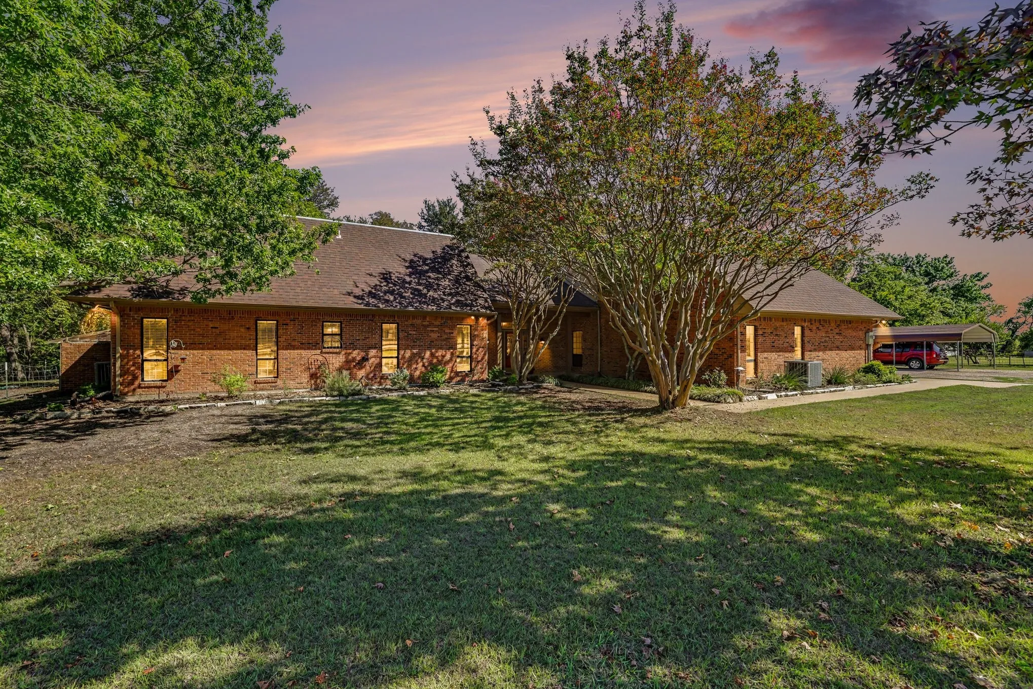 Brick exterior, gabled roofline, multiple windows, established trees, and a covered carport
