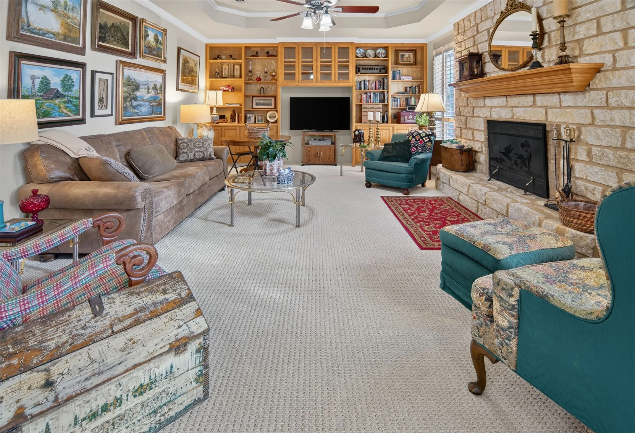 Recessed ceiling with a ceiling fan, built-in wooden shelving, a stone fireplace, and light-toned carpet flooring