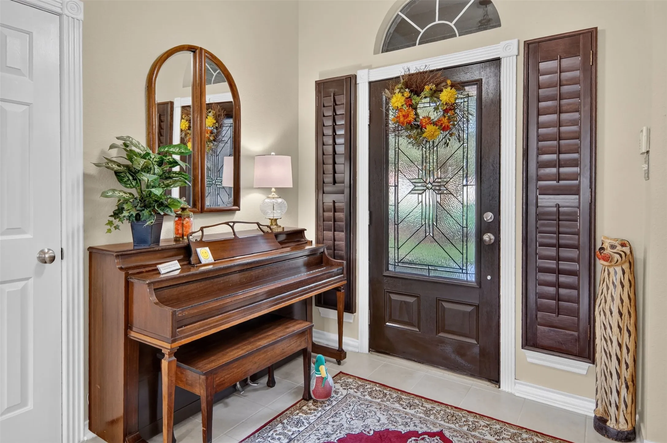 Transom window with decorative grille, entry door with leaded glass insert, wood-finish interior shutters, and tile flooring