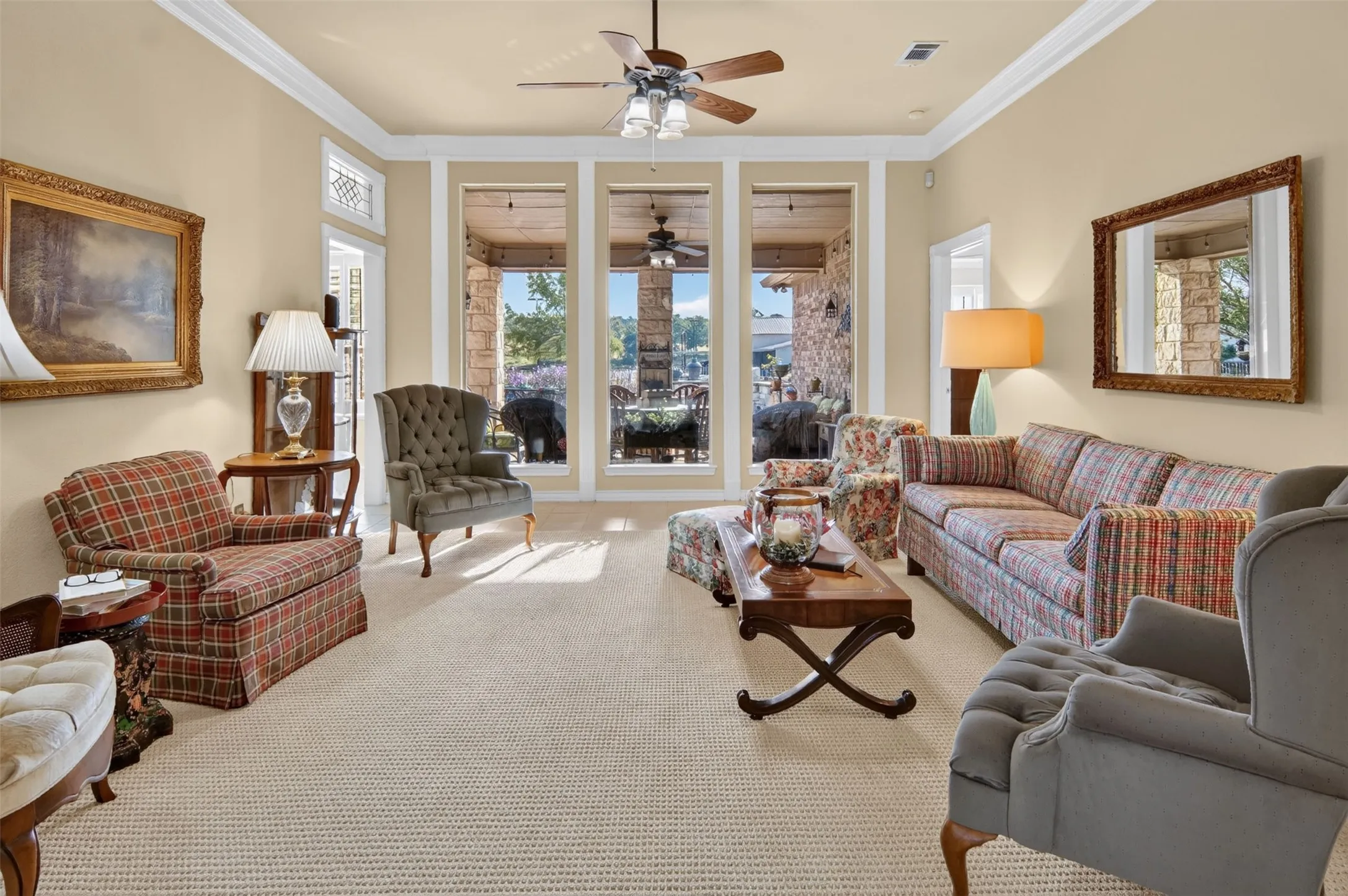 Tray ceiling with crown molding, carpeted flooring, three large windows, outdoor covered patio with stone accents, and a ceiling fan