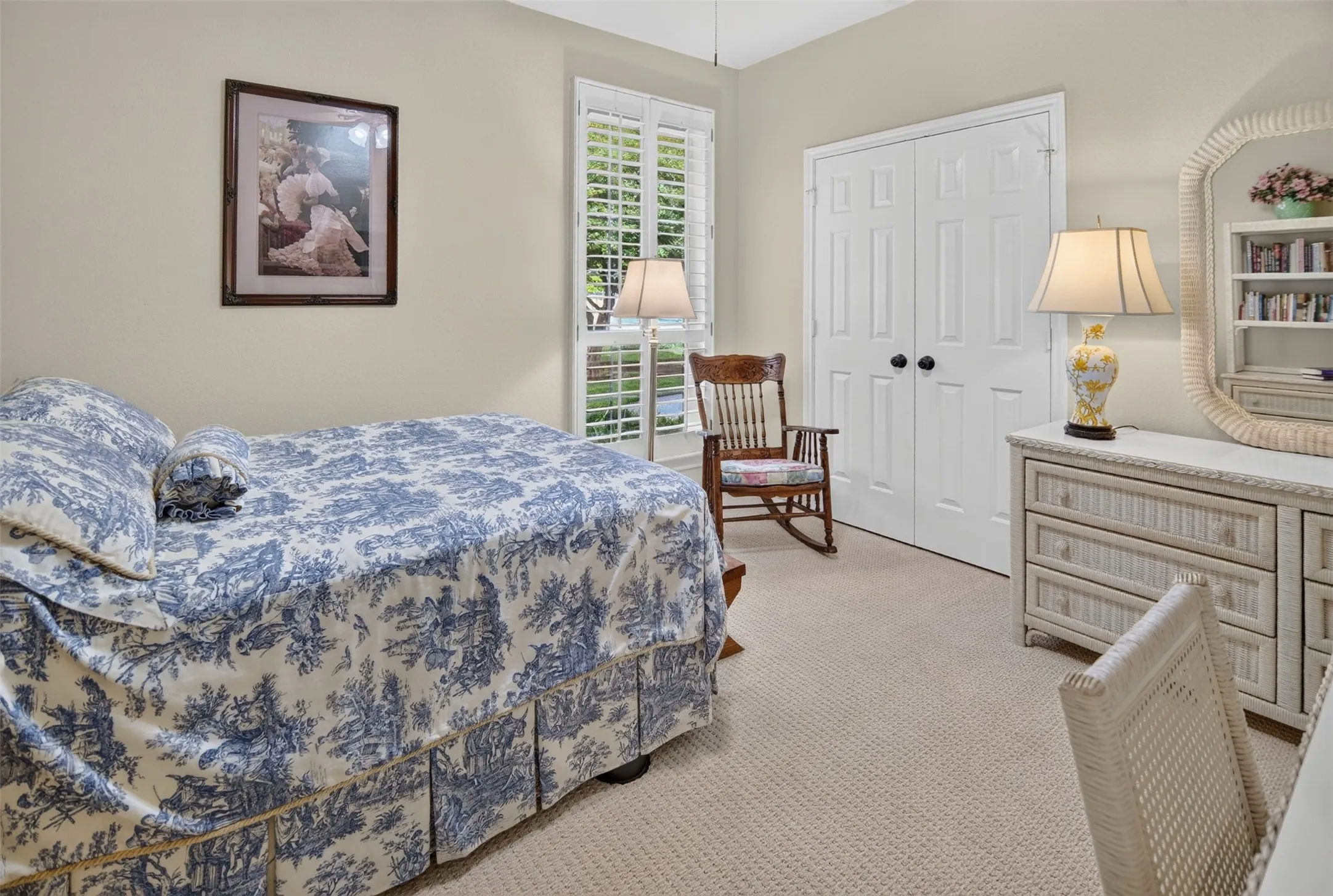 Serene bedroom retreat featuring a blue toile bedspread, cream carpeting, white double-door closet, decorative mirror, wood-finish dresser with attached lighting, and shuttered French doors opening to natural light