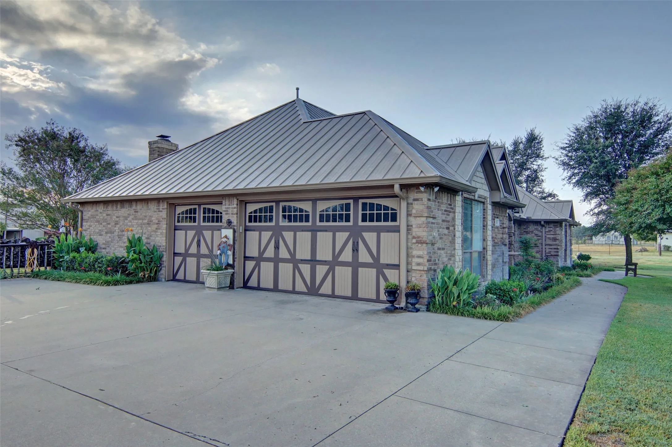 Stone veneer exterior with a metal roof, two-car garage with decorative doors, concrete driveway, and established landscaping