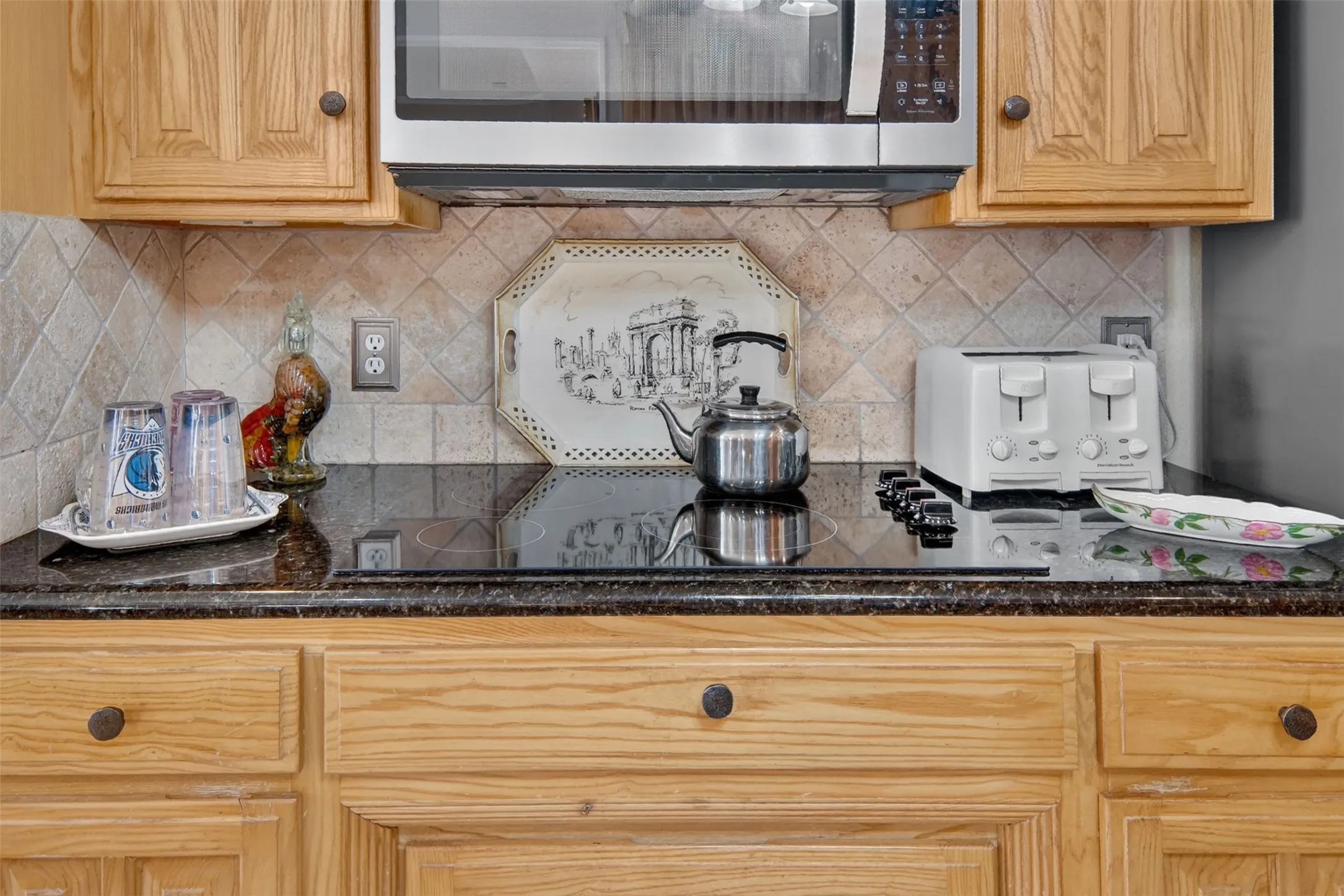 Wood-finish cabinetry, dark speckled countertop, diamond-patterned tile backsplash, stainless steel microwave, and a black cooktop