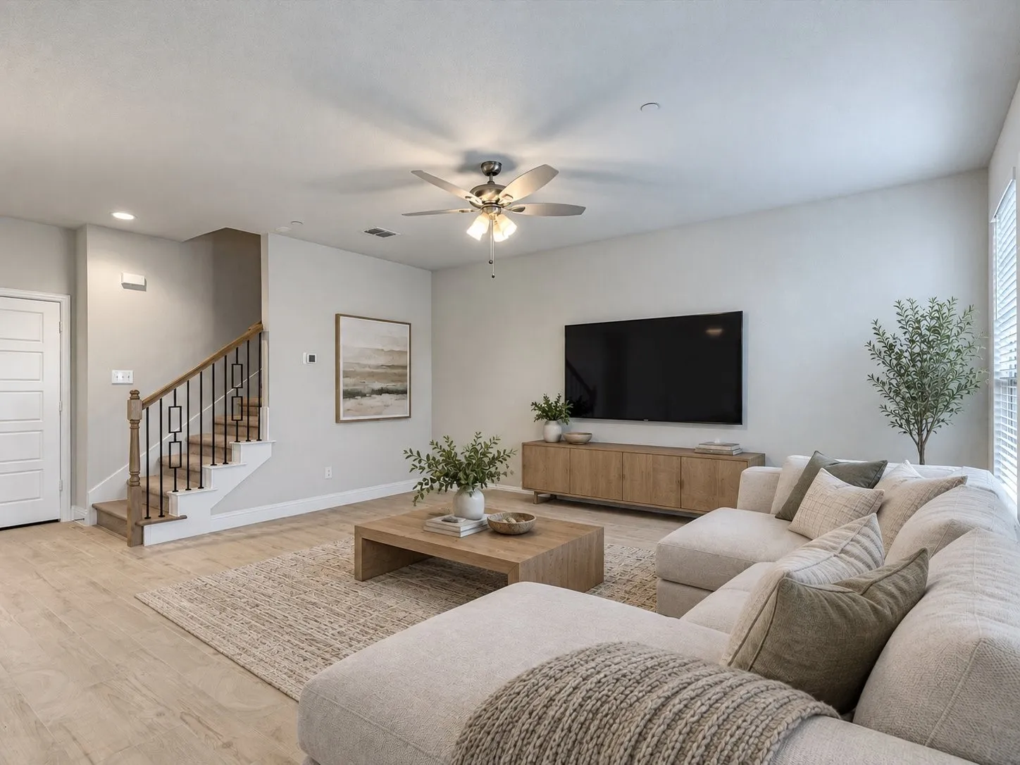 Light-toned wood-finish flooring, staircase with wood handrail and iron balusters, ceiling fan with integrated lighting, and white paneled door