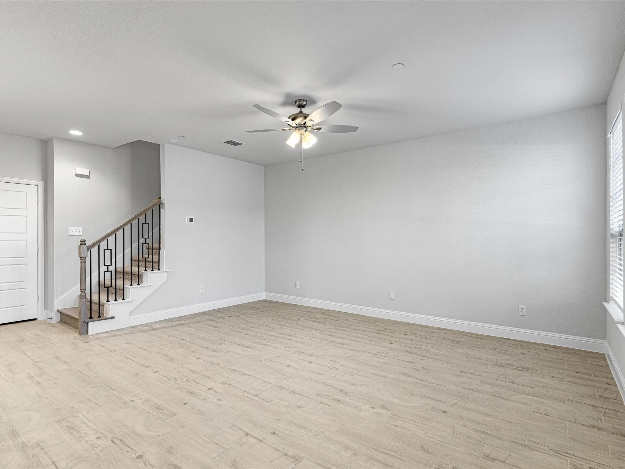 Spacious room featuring wood-finish flooring, a contemporary staircase with iron balusters, a ceiling fan with integrated lighting, and natural light from a window