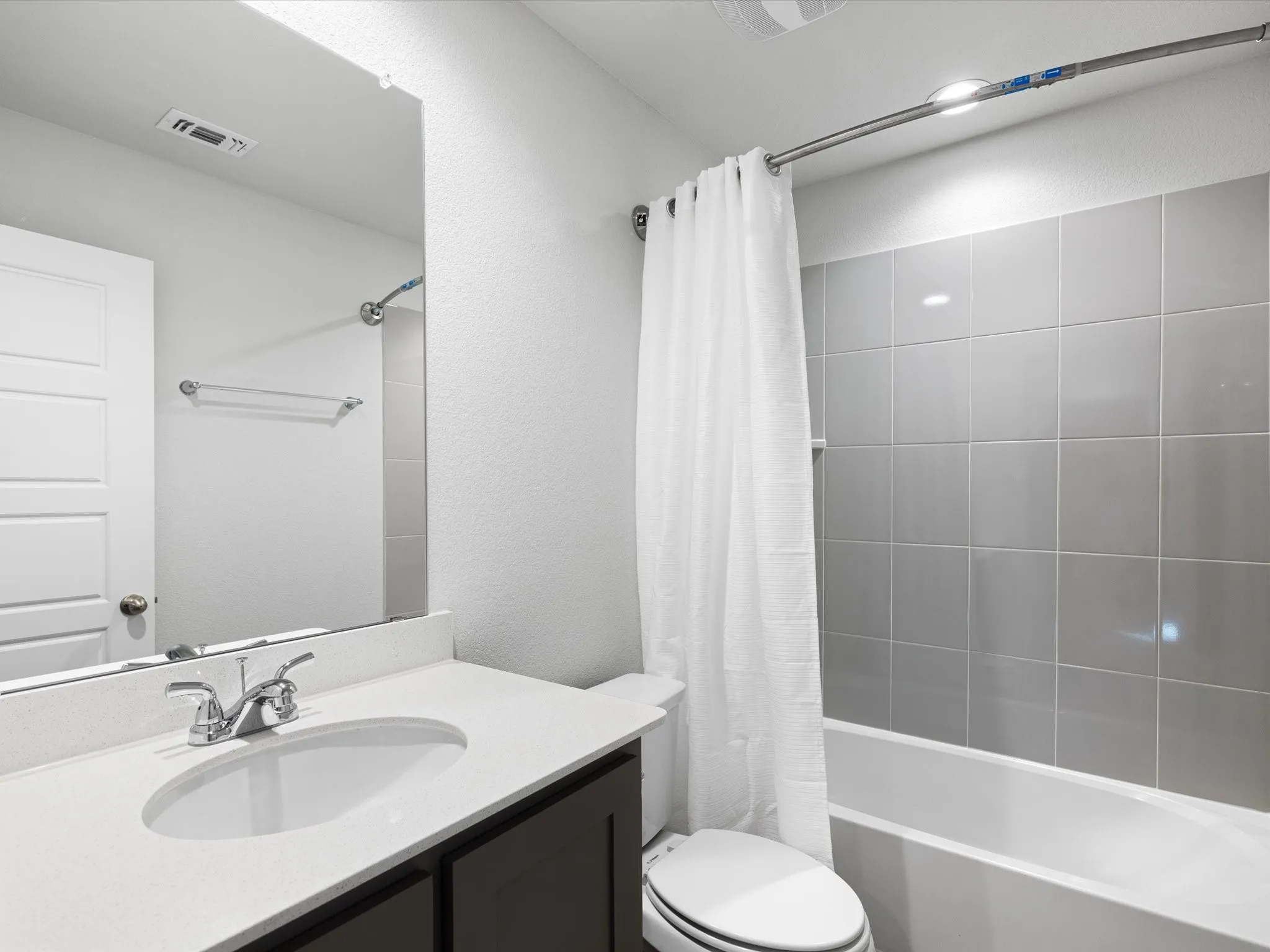 White bathtub with grey tile surround, dark wood-finish vanity with a white countertop, and a large wall-mounted mirror