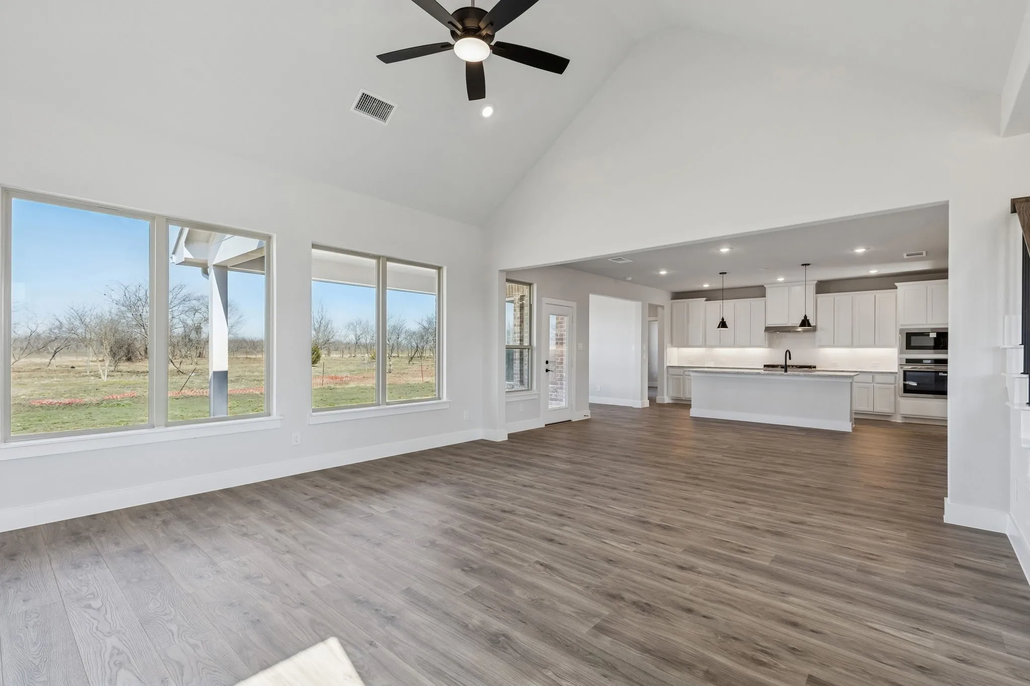 Vaulted ceiling with a ceiling fan, wood-finish flooring, ample natural light from large windows, and a kitchen featuring a central island and white cabinetry